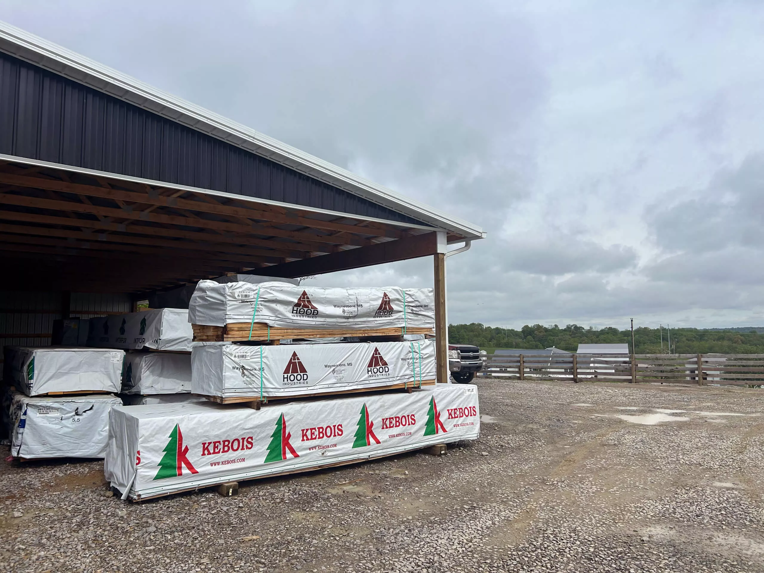 Amish Ridge Metals Perry, OH Stacks of lumber wrapped in plastic labeled "Hood" and "Kebois" are stored beside a large metal-roofed building on a gravel lot under a cloudy sky.