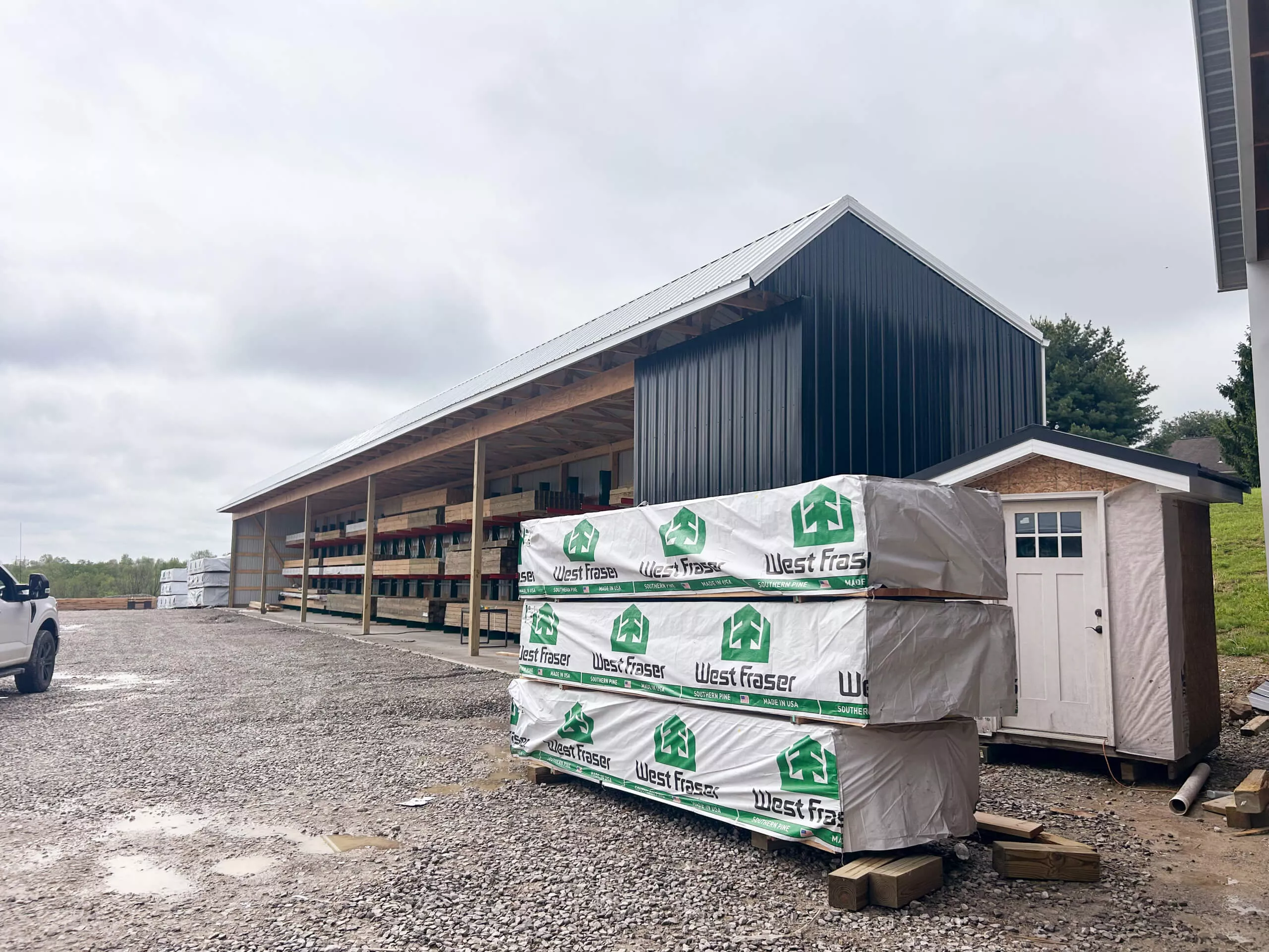 Amish Ridge Metals Perry, OH A large metal building with open sides stands beside stacks of lumber wrapped in West Fraser packaging; a small shed is positioned nearby on a gravel lot.