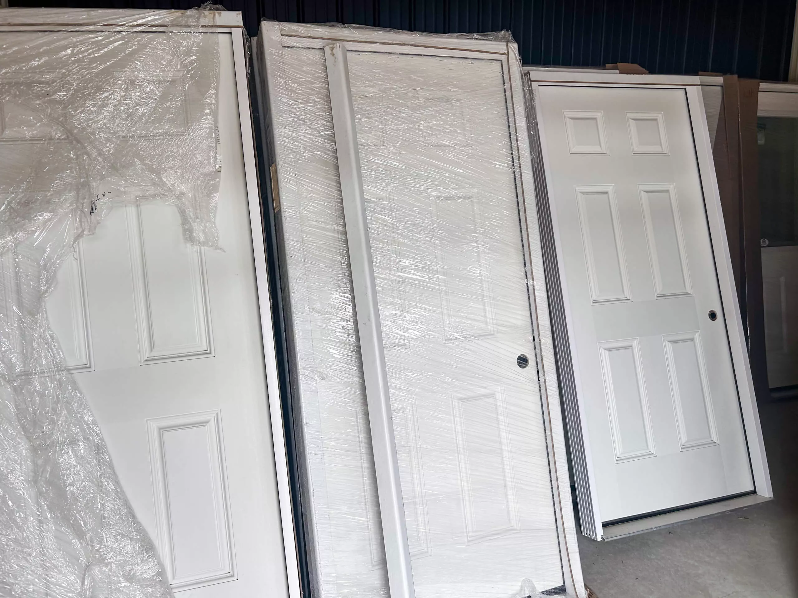 Amish Ridge Metals Perry, OH Several white panel doors, some wrapped in plastic and some unwrapped, are standing upright against a wall in what appears to be a storage or warehouse area.