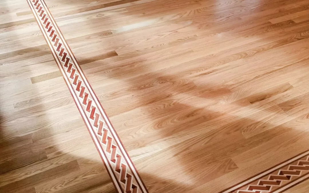 Cove Mountain Wood Products Bradford, PA Hardwood floor with a decorative inlaid border featuring a geometric pattern, located in a room with a stone fireplace and green walls.