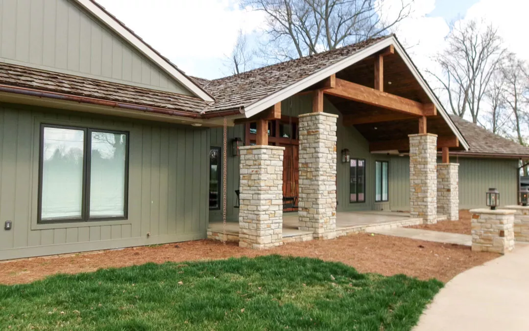 Miller's Guttering Seamless Gutters Arthur, IL A single-story house with green siding, large windows, and a covered front porch supported by stone columns. The lawn is partially green with some bare soil areas.