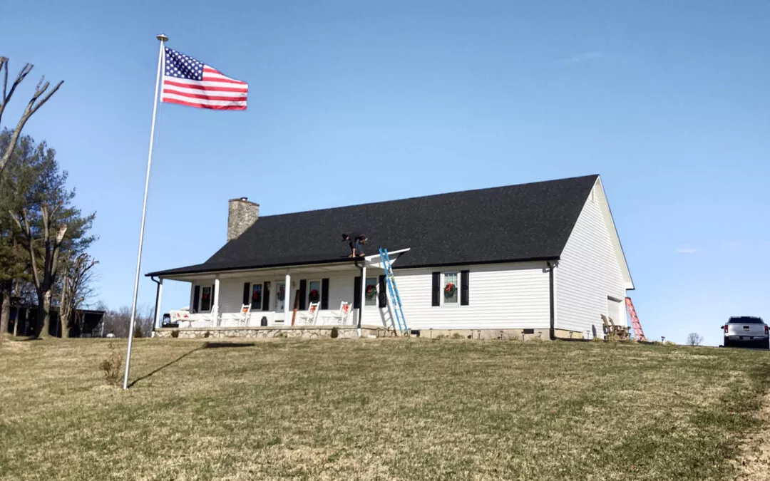 Miller's Guttering Seamless Gutters Arthur, IL A single-story white house with a black roof sits on a grassy slope. An American flag is raised on a flagpole, and a person works on the roof using a blue ladder.