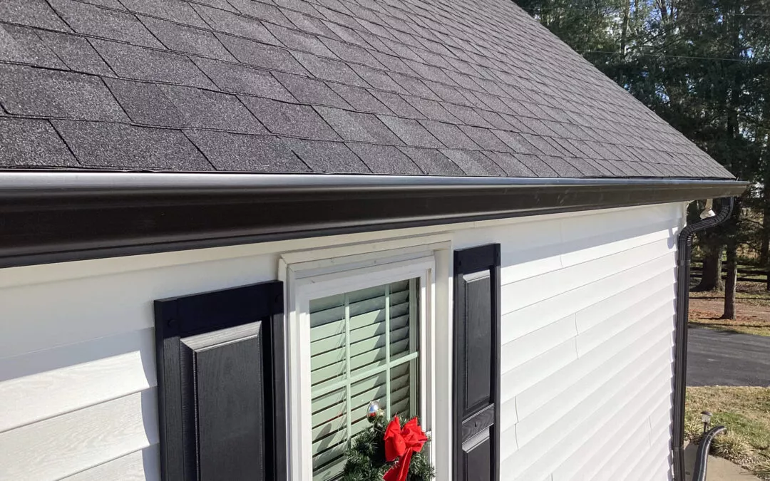 Miller's Guttering Seamless Gutters Arthur, IL A close-up of a house exterior with black roof shingles, black gutters, white siding, and black window shutters. A window with blinds has a wreath with a red bow hanging on it.