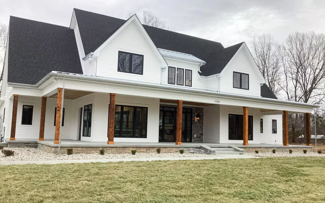 Miller's Guttering Seamless Gutters Arthur, IL A large two-story white house with black roof shingles, wooden pillars, and a wraparound porch, set on a grassy lawn with a stone border and leafless trees in the background.