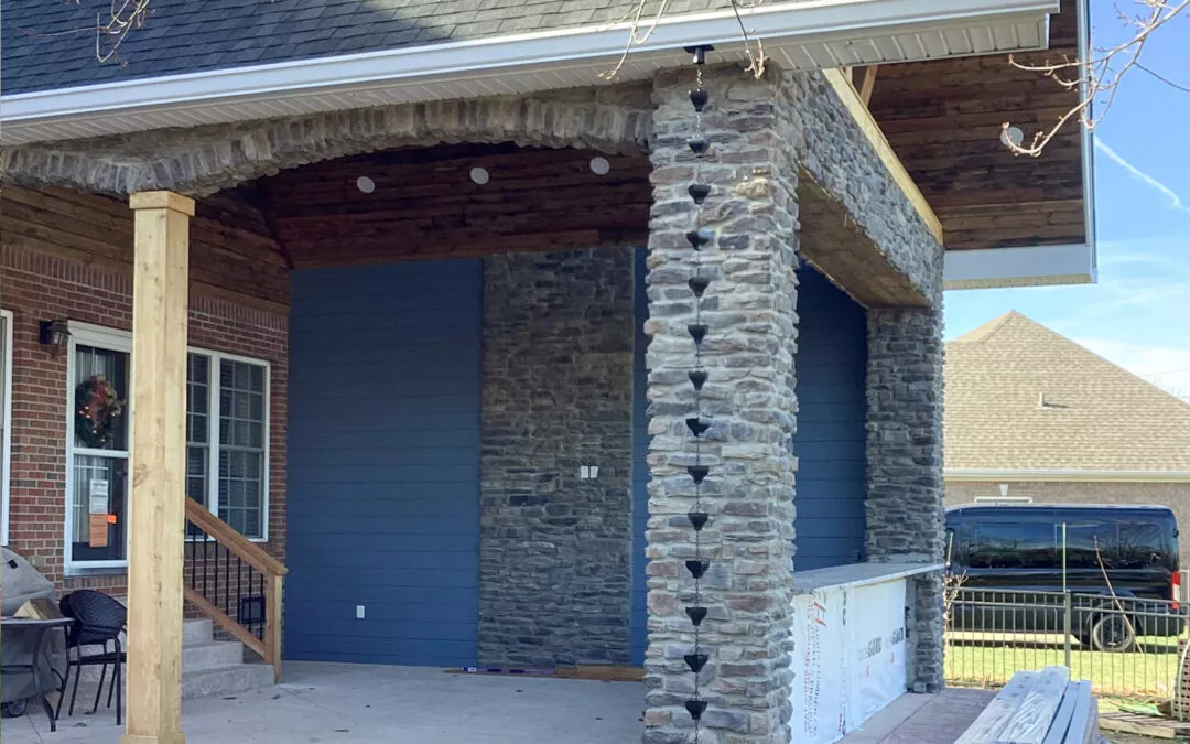 Miller's Guttering Seamless Gutters Arthur, IL A covered patio area with stone pillars, wood ceiling, blue siding, a rain chain, and construction materials on a counter under the roof.