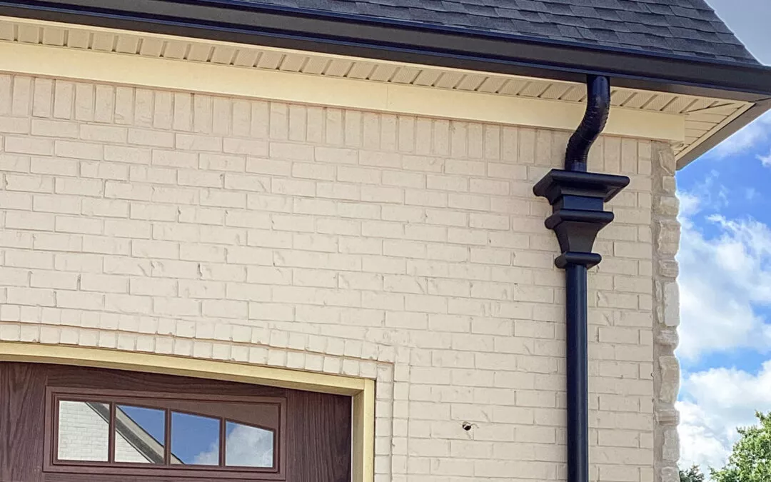 Miller's Guttering Seamless Gutters Arthur, IL Black metal downspout with decorative bracket attached to the light brick exterior of a building next to a wooden garage door under a cloudy sky.