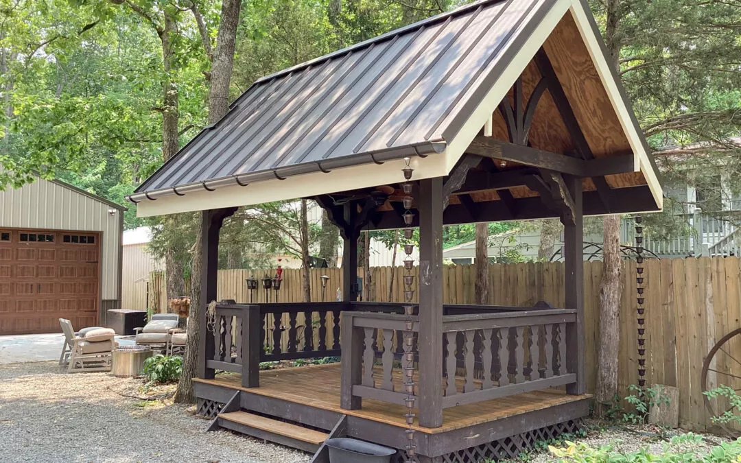 Miller's Guttering Seamless Gutters Arthur, IL A wooden gazebo with a peaked metal roof stands on a gravel area next to a wooden fence, surrounded by trees, with outdoor furniture nearby.