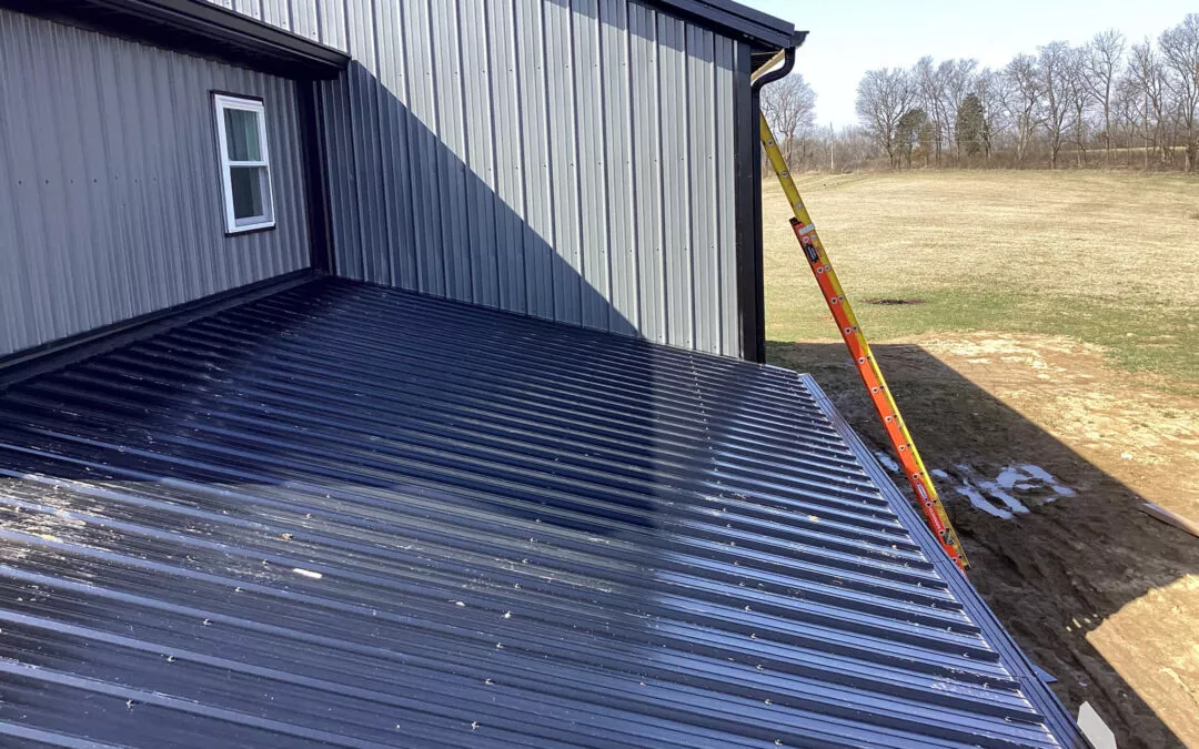 Miller's Guttering Seamless Gutters Arthur, IL A metal roof on a building with a closed window, a ladder resting against the gutter, and an open field with trees in the background.