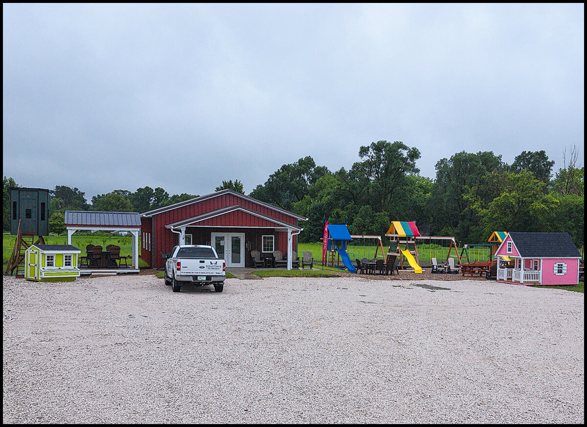 7.12.25 Backyard & Beyond JPB Summary Graphic Right A gravel lot with a white truck parked in front of a red building, surrounded by colorful playground equipment and small playhouses under a cloudy sky.