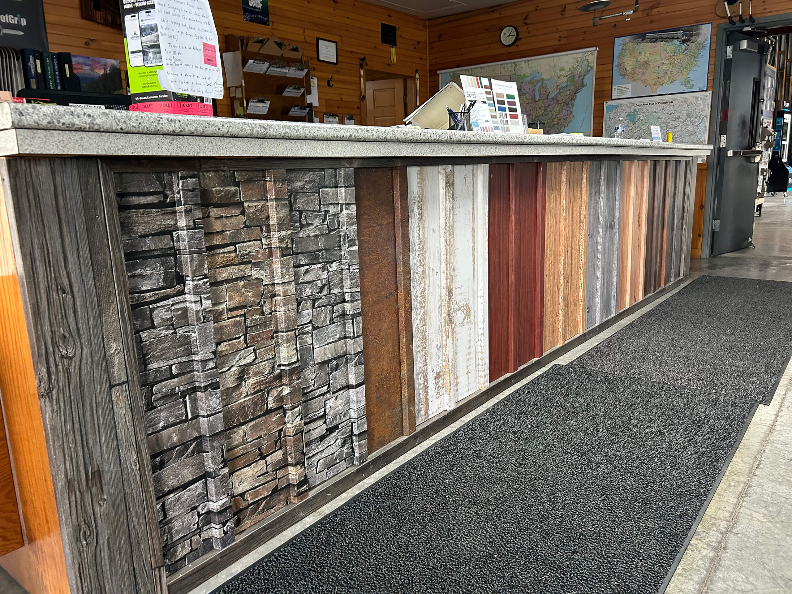 Dave's Metal Roofing Clare MI A front desk with a countertop and a facade decorated with various wood and stone-patterned panels, located in a room with maps and signs on the wall.