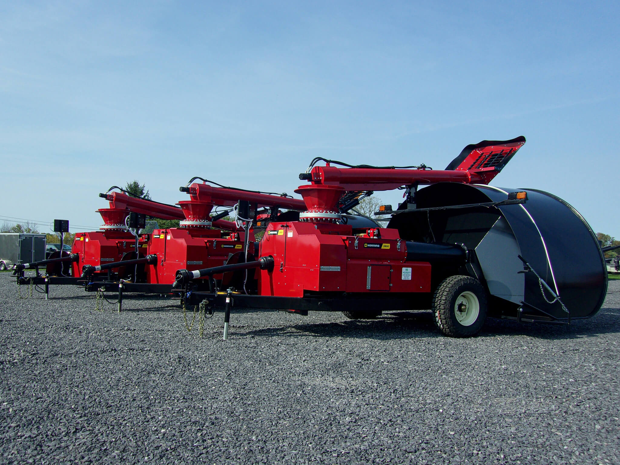 Three large red grain augers with black hoppers are parked in a row on a gravel lot under a clear sky.