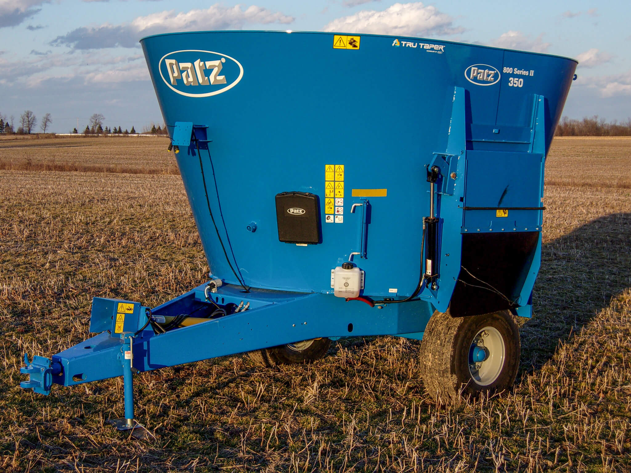 A blue Patz 350 feed mixer is parked on a harvested field under a partly cloudy sky.