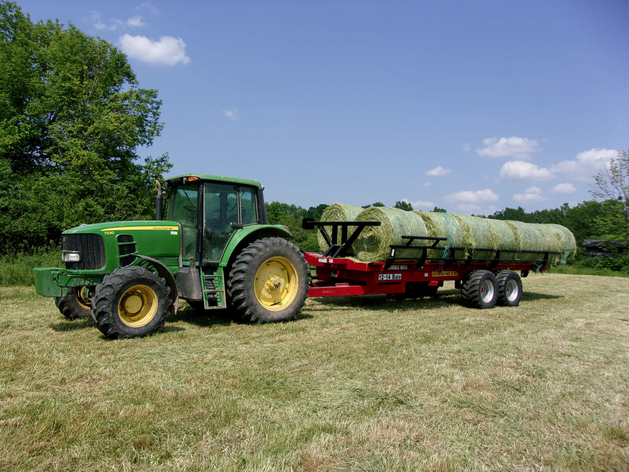 A green tractor is pulling a red trailer loaded with large round hay bales across a grassy field under a clear sky.