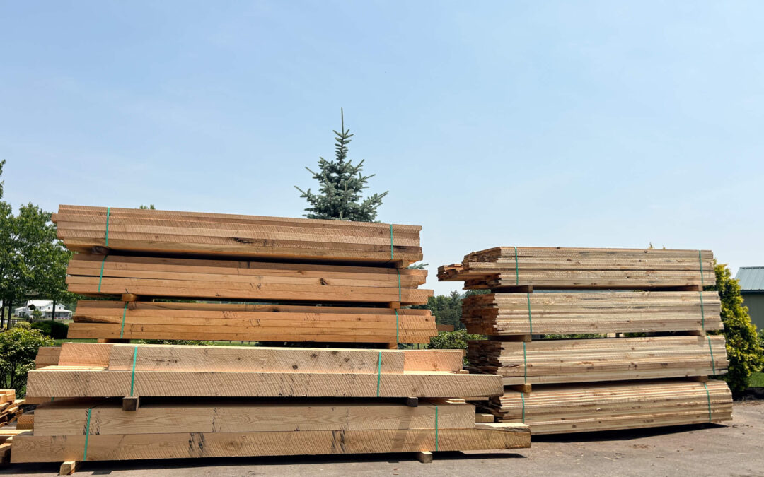 Leinbach's Sawmill Lumber Lancaster, PA Stacks of large wooden beams and planks bundled with green straps, placed outdoors on a paved surface with trees in the background.