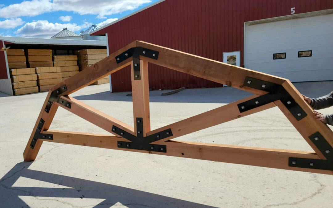 Martin's Truss Manufacturing Elkhart IN A large wooden roof truss with black metal brackets is held outdoors near a red building and stacks of lumber under a partly cloudy sky.