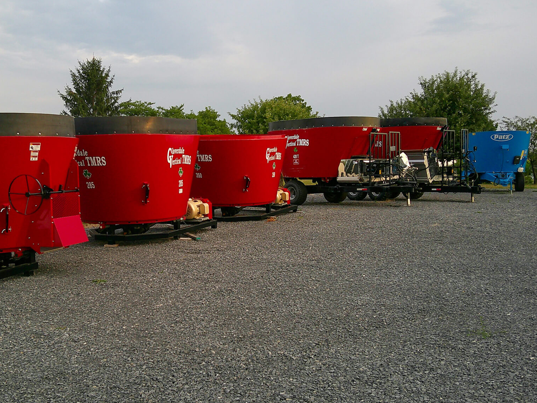 Several red agricultural feed mixer wagons and one blue wagon are parked on a gravel lot with trees and sky in the background.