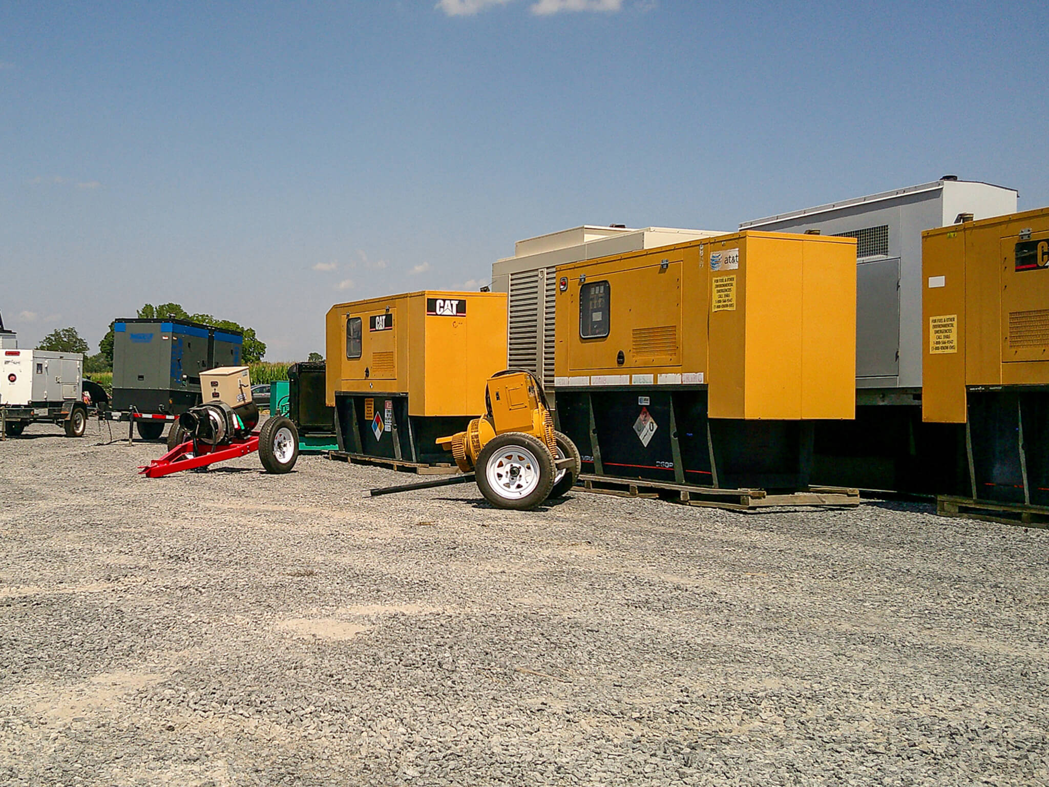 Several large yellow industrial generators are lined up on a gravel lot under a clear blue sky, with a few other trailers and equipment visible in the background.