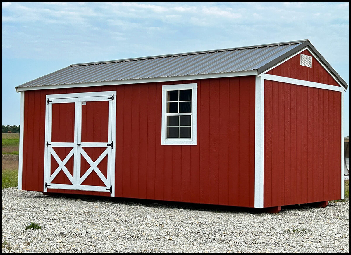 Gingerich Portable Buildings Jamesport MO A red wooden shed with a gray metal roof, white trim, double doors, and a single window, standing on a gravel surface.