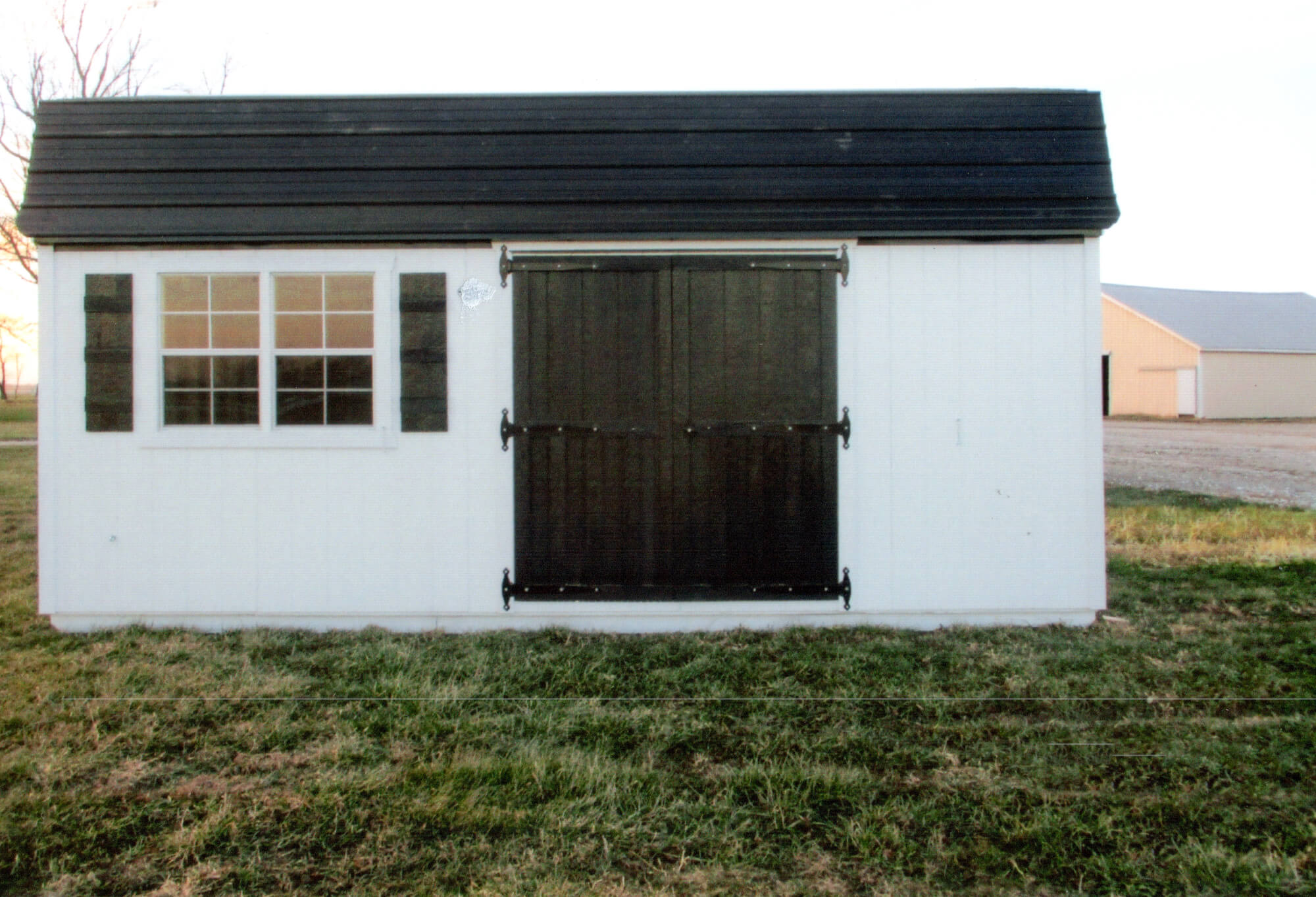 Woodcraft Storage Buildings Boone IA A small white shed with a black roof, double barn doors, and a window with shutters stands on grass with another building visible in the background.