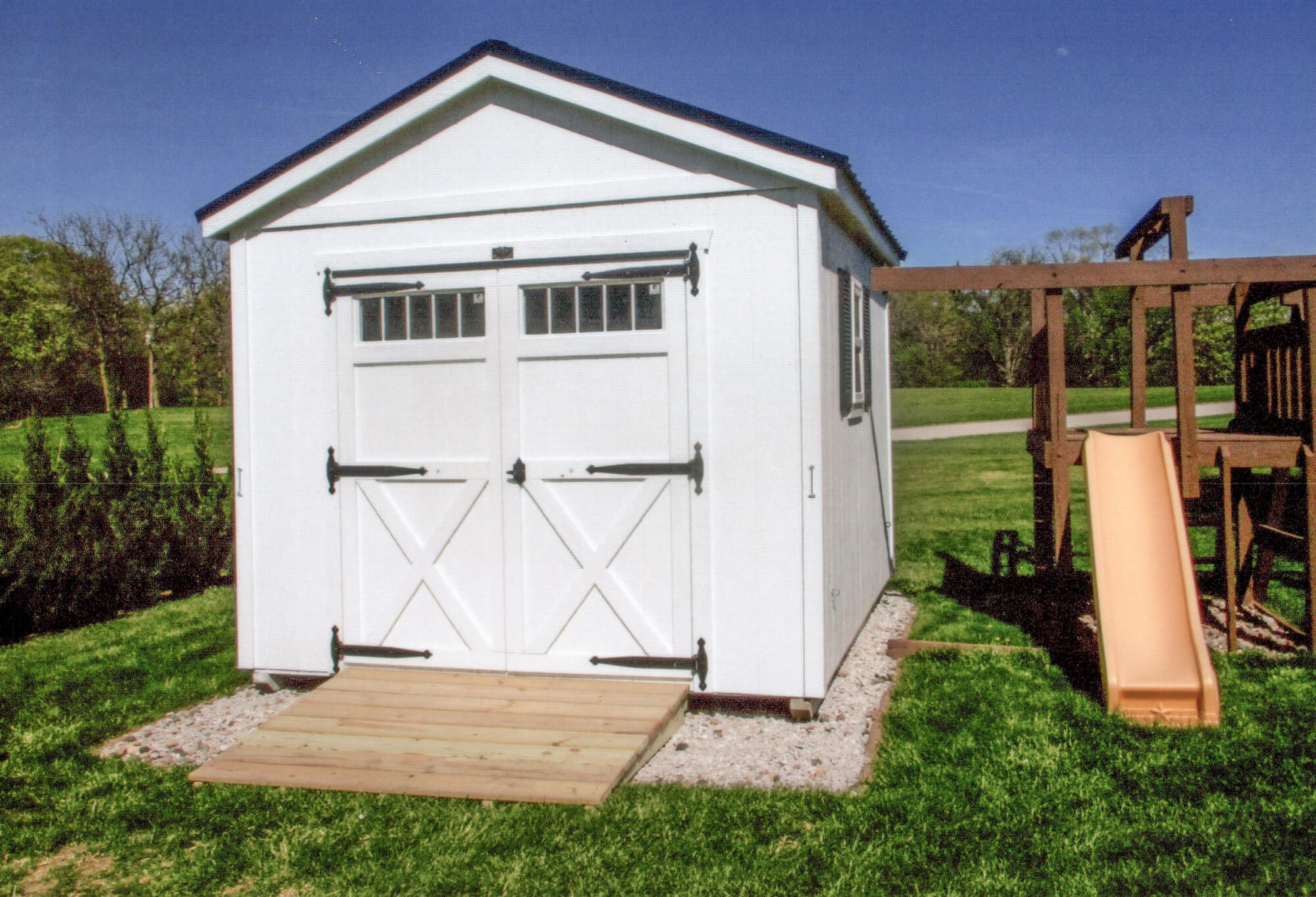 Woodcraft Storage Buildings Boone IA A white storage shed with double doors and a ramp, next to a wooden playset with a yellow slide, on a grassy lawn.