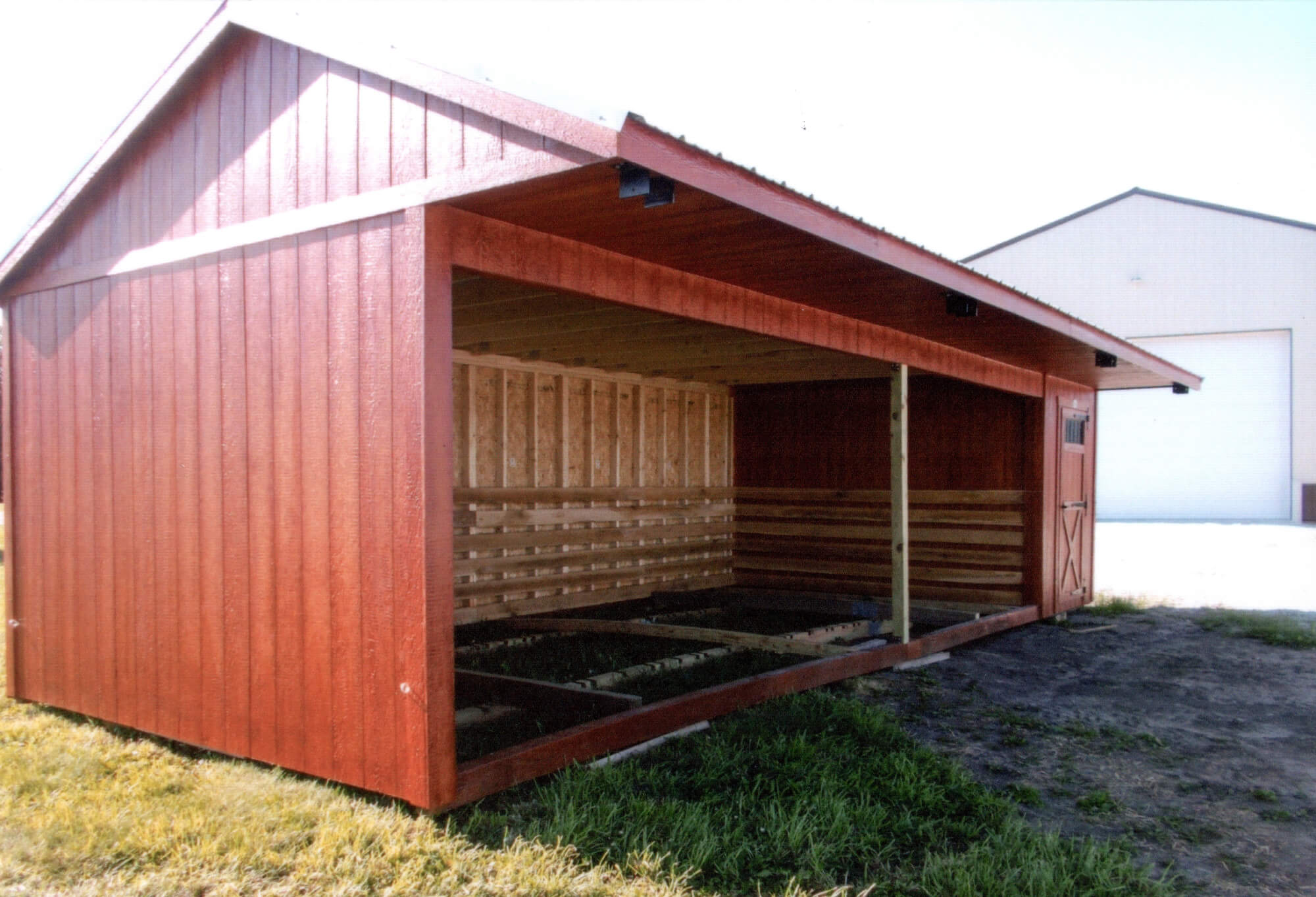 Woodcraft Storage Buildings Boone IA A red wooden open-sided shed with a gabled roof and a small closed door on the right side, standing on grass near another building.