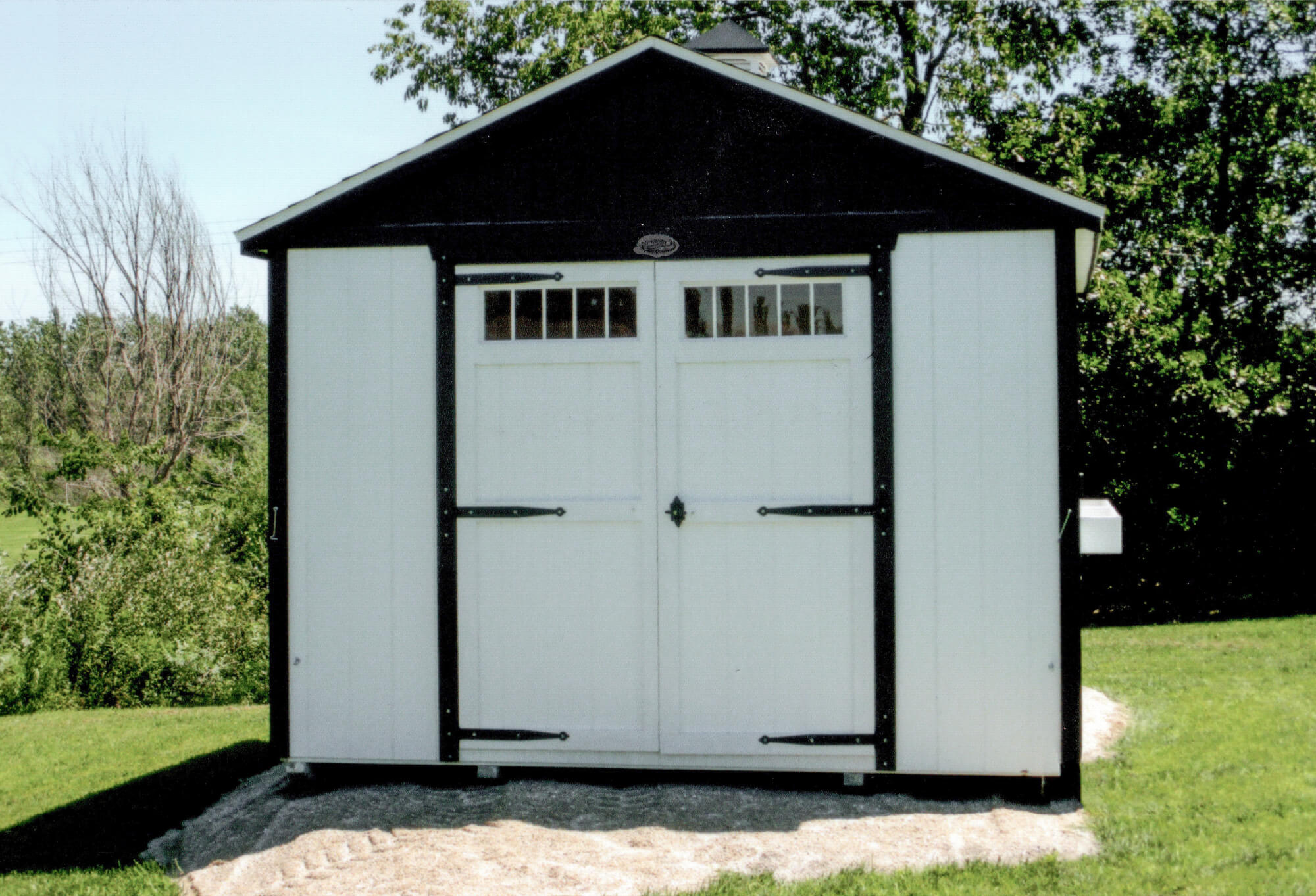 Woodcraft Storage Buildings Boone IA A small white storage shed with double doors and black trim sits on a grassy lawn with trees in the background.