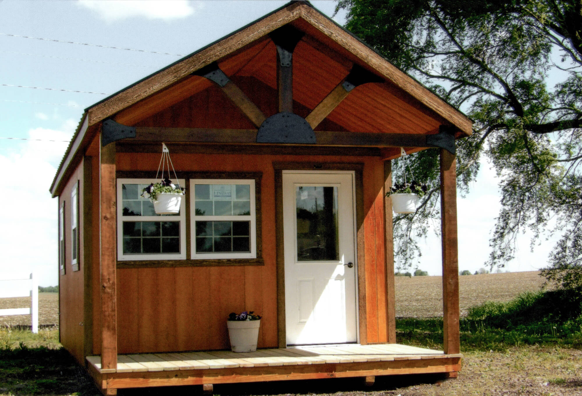Woodcraft Storage Buildings Boone IA Small wooden shed with a covered porch, white door, two windows, hanging flower baskets, and a potted plant on the steps; situated in a rural outdoor setting.
