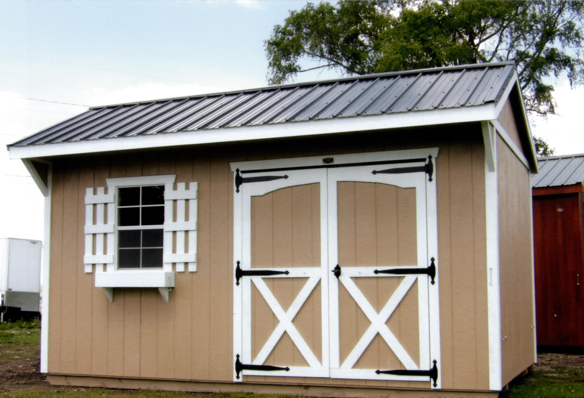 Woodcraft Storage Buildings Boone IA A tan storage shed with a metal roof, double white doors with black hardware, and a window with white trim and shutters, placed outdoors on a grassy area.