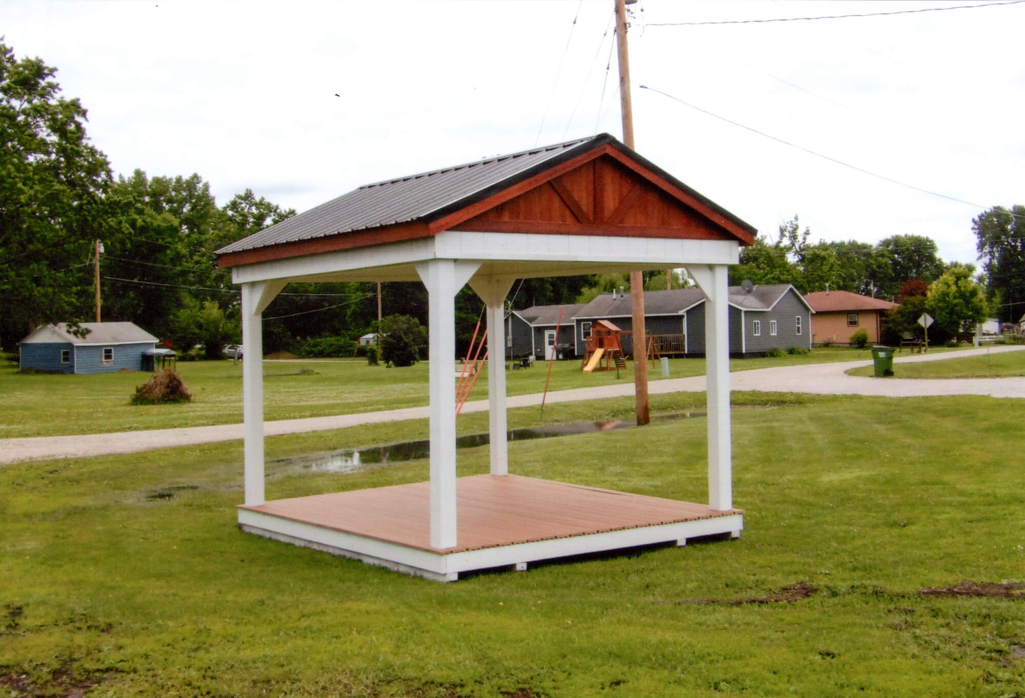 Woodcraft Storage Buildings Boone IA A small, open wooden pavilion with a gable roof stands on a grassy lawn near a residential area with houses and a playground in the background.