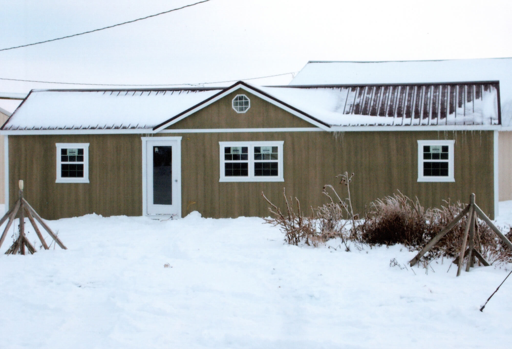 Woodcraft Storage Buildings Boone IA A single-story tan building with three windows and a door, covered in snow with bare bushes and wooden supports in the snowy foreground.