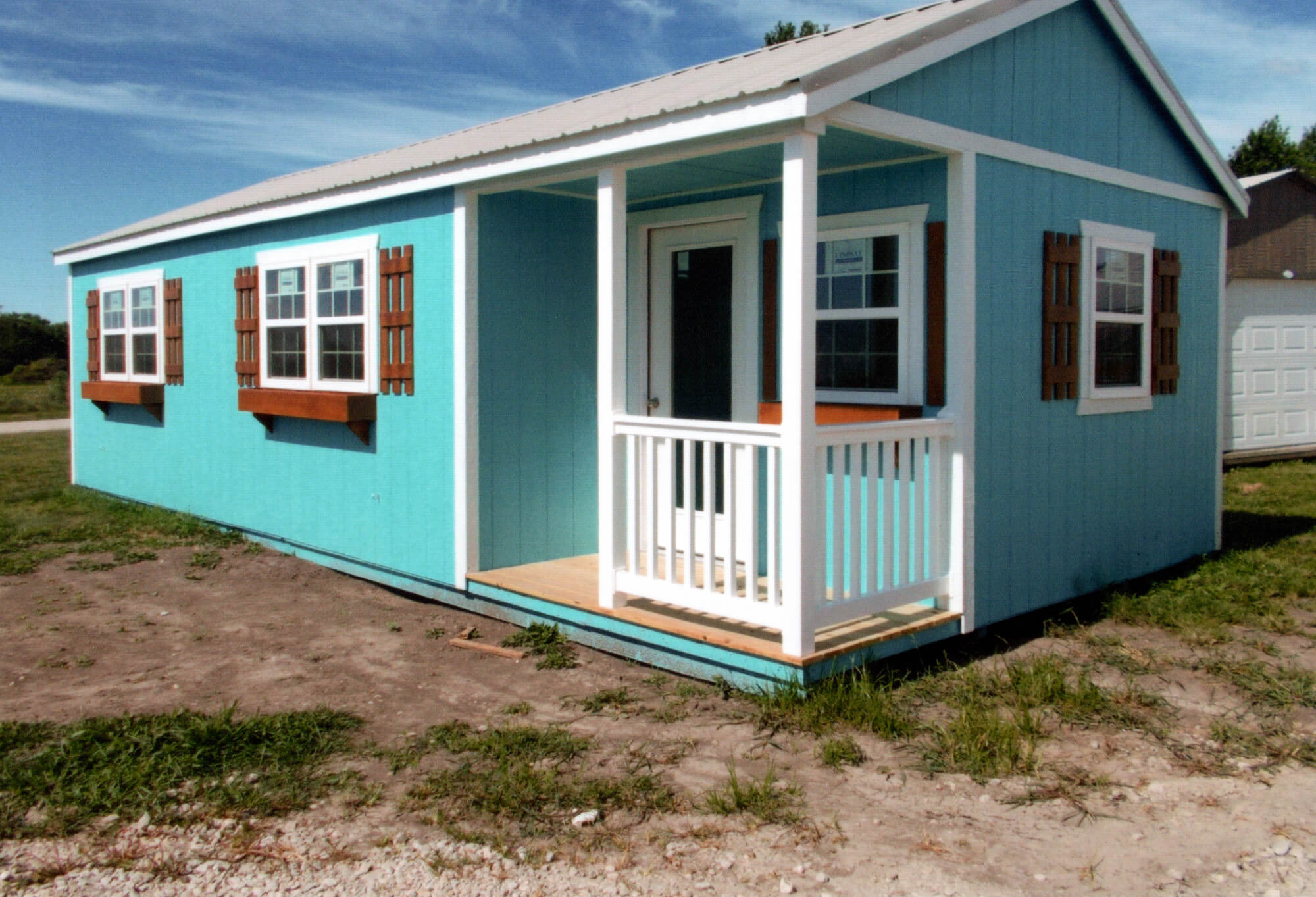 Woodcraft Storage Buildings Boone IA A small, single-story blue house with white trim, a front porch, and brown window shutters sits on a patch of grass and dirt under a blue sky.