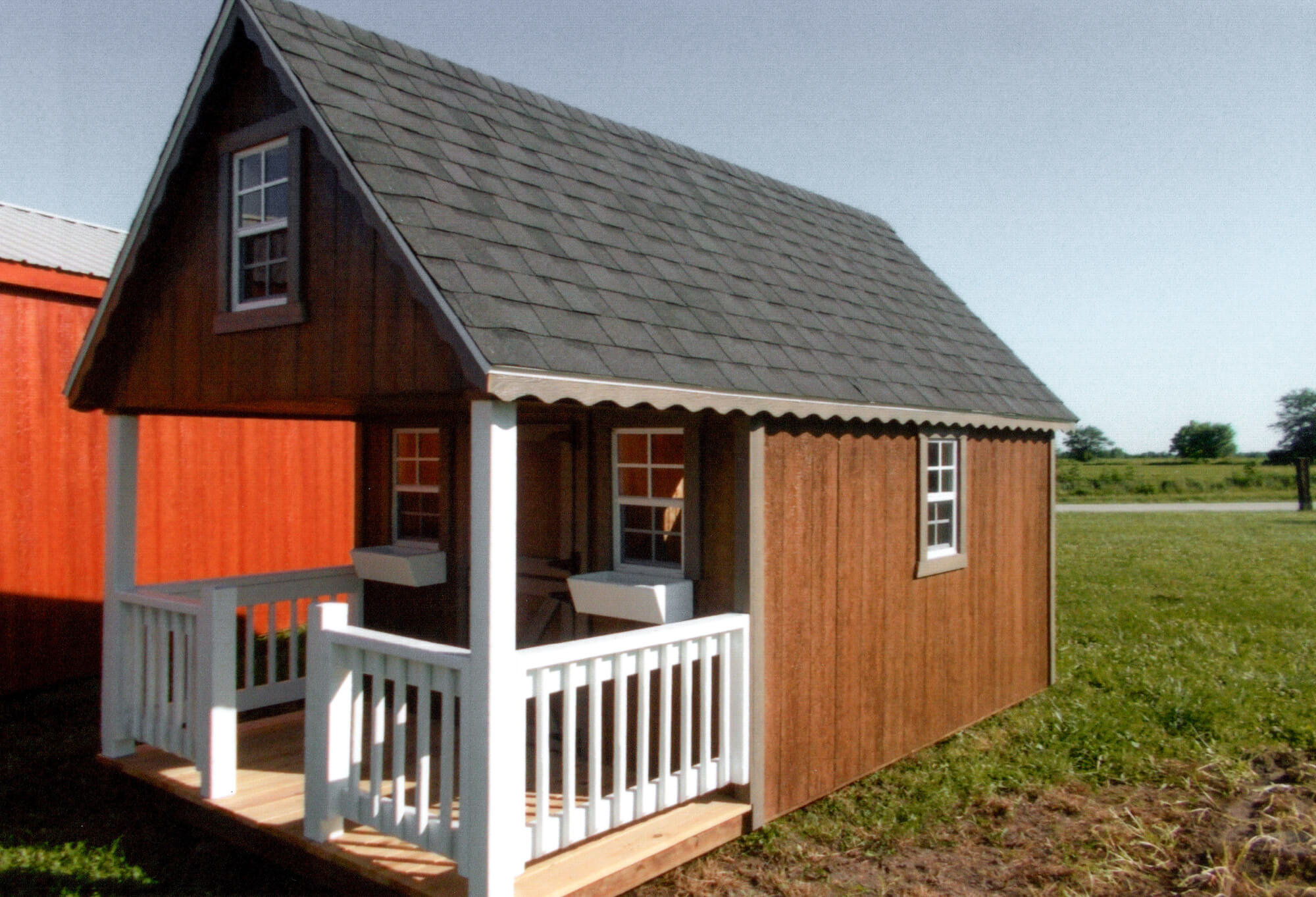 Woodcraft Storage Buildings Boone IA Small wooden playhouse with a gray shingle roof, porch, and white railing, set on grass near a red building and open field under a clear sky.