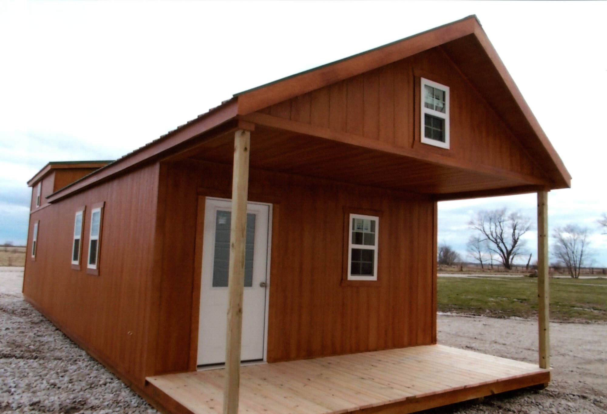Woodcraft Storage Buildings Boone IA A small wooden house with a covered front porch, three visible windows, and a white door, situated on a gravel surface with grass and bare trees in the background.