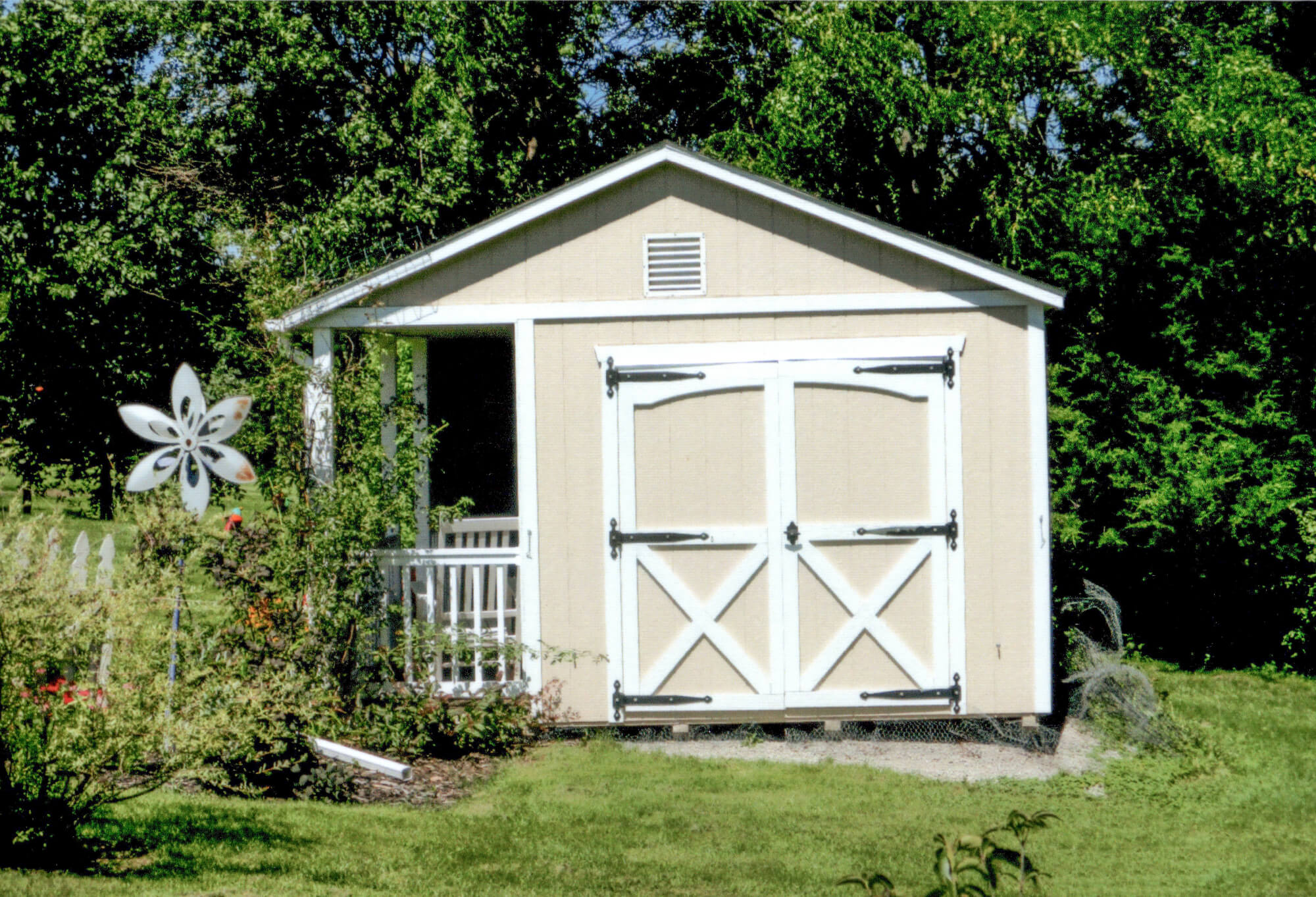 Woodcraft Storage Buildings Boone IA A beige garden shed with white double doors and black hinges, surrounded by greenery with a windmill decoration to the left.