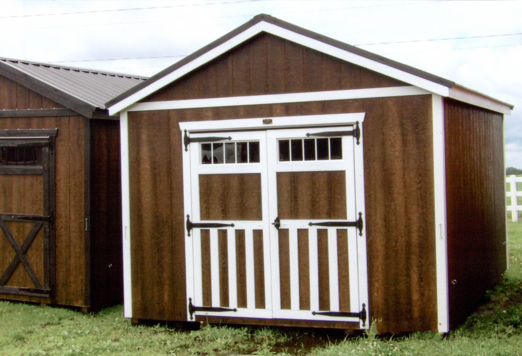 Woodcraft Storage Buildings Boone IA A brown wooden storage shed with a white-trimmed double door and windows, situated on grass next to another similar structure.