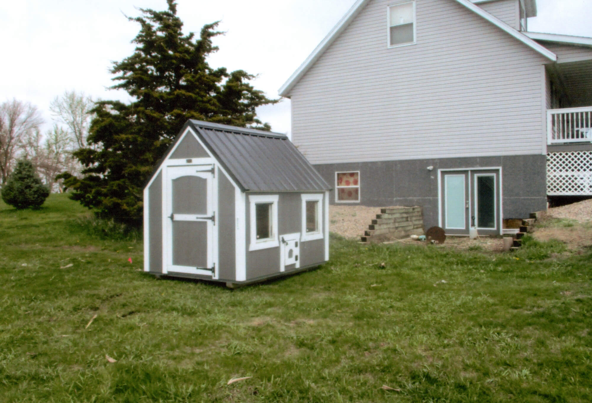 Woodcraft Storage Buildings Boone IA A small gray and white shed with windows sits on grass near a house with gray siding and a raised porch.