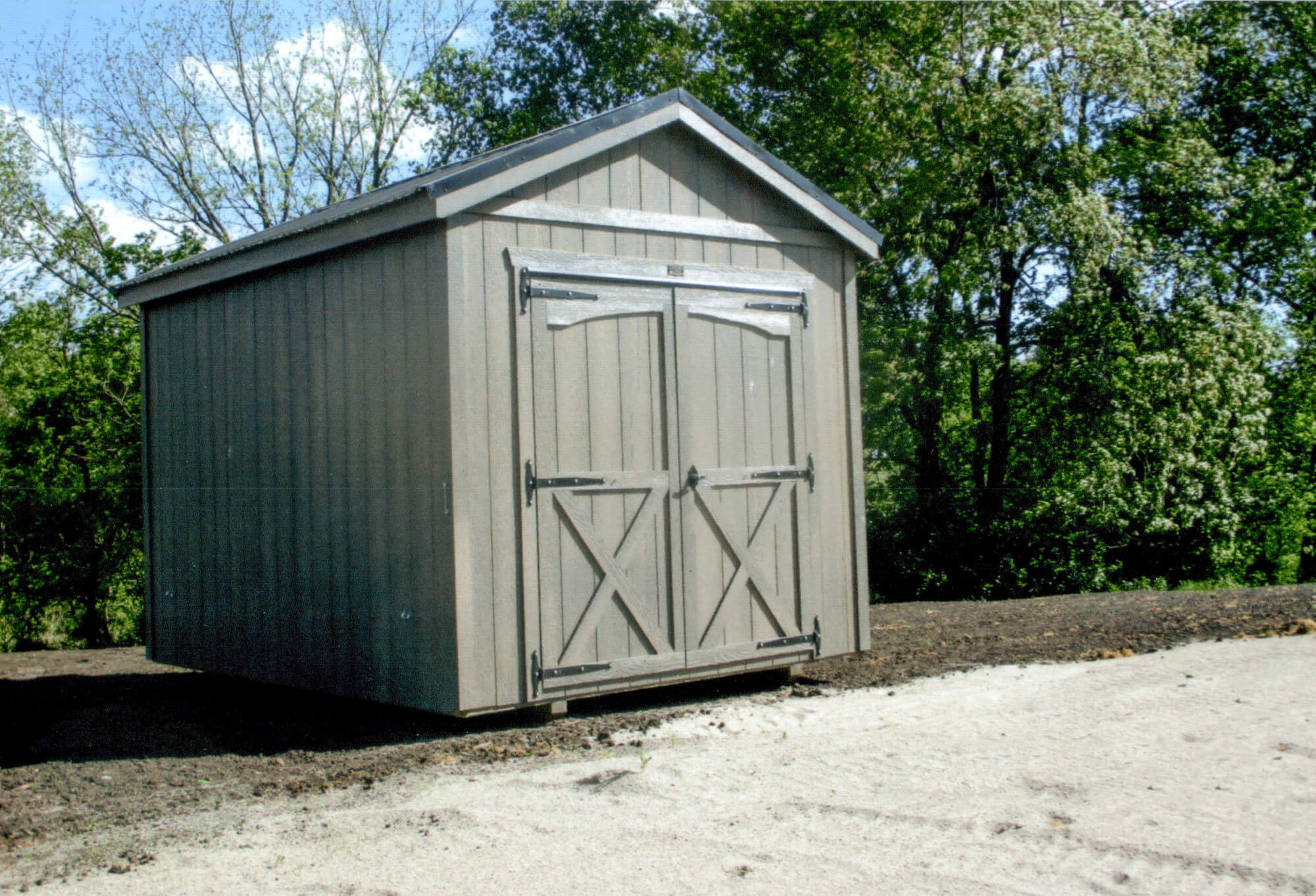 Woodcraft Storage Buildings Boone IA A small, wooden shed with double doors sits on bare ground, surrounded by trees and greenery in the background.