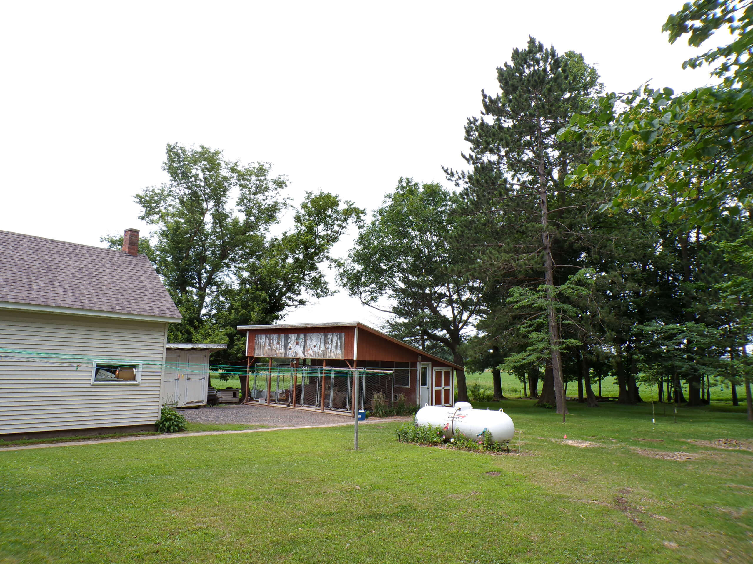 A backyard with a lawn, a tan house, a screened porch, two white propane tanks, and several large trees in the background.