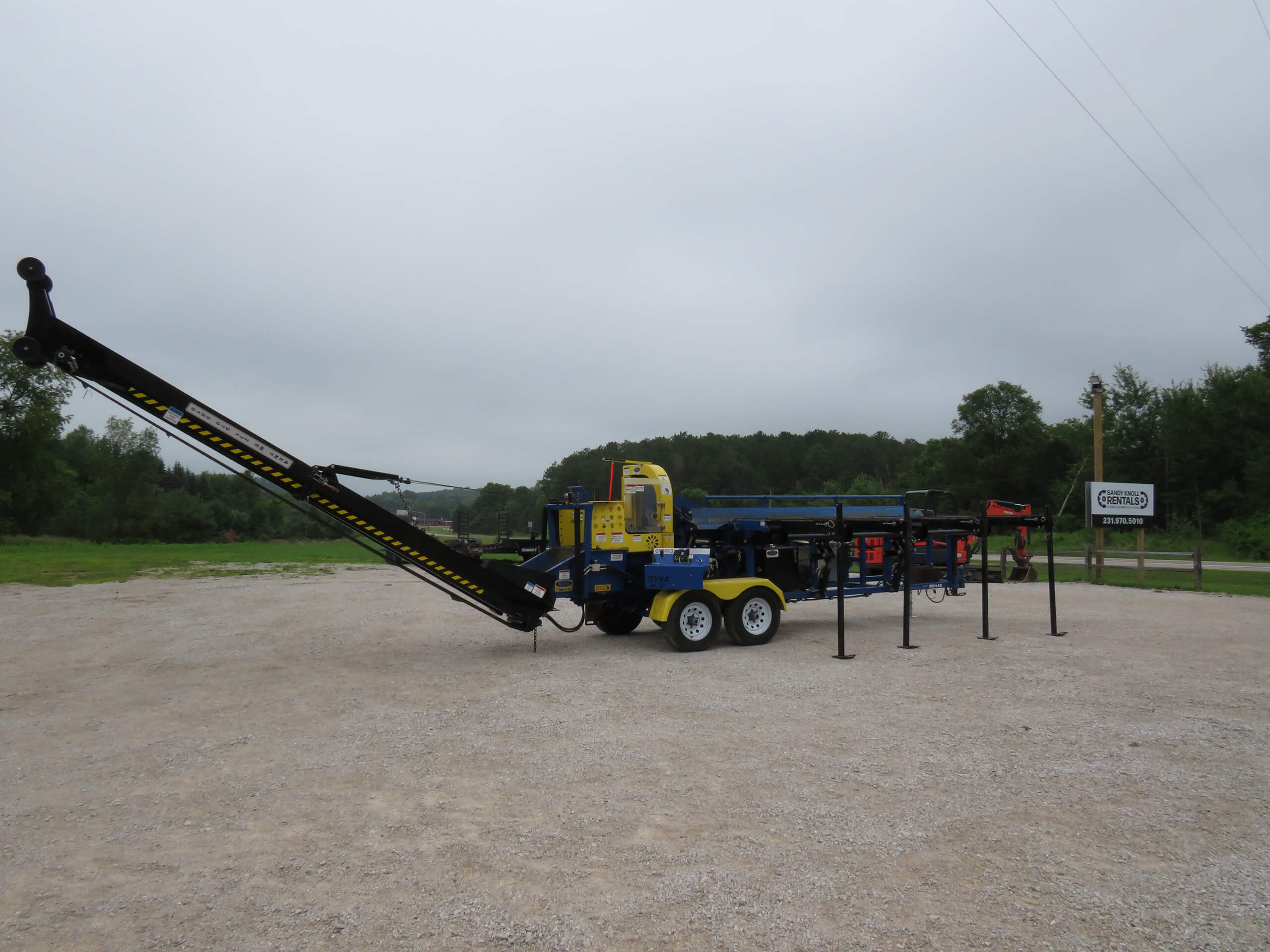 A large yellow and blue industrial machine with a conveyor arm is parked on a gravel lot surrounded by trees under a cloudy sky.
