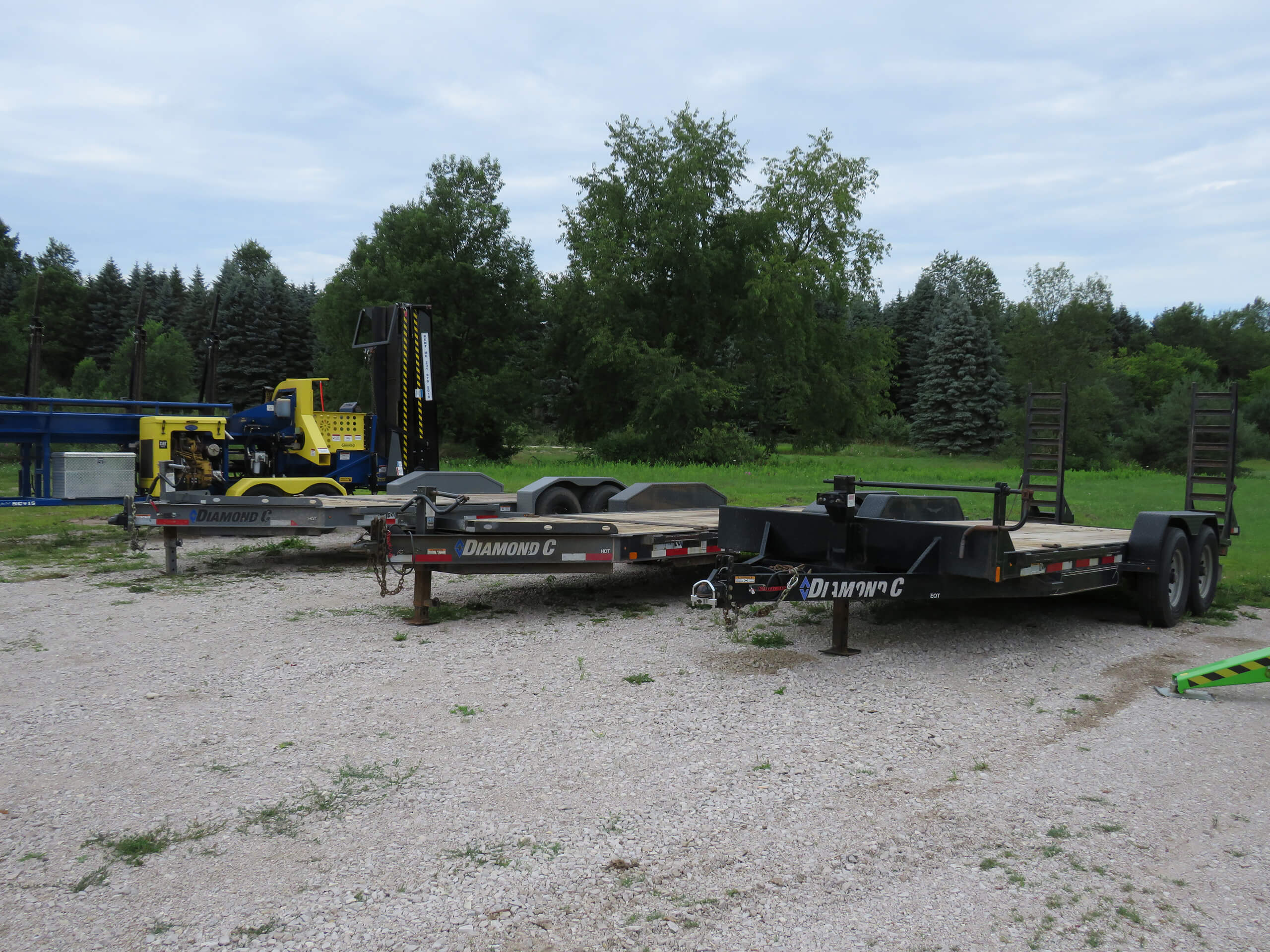 Two Diamond C trailers are parked on a gravel surface near a yellow construction vehicle, with trees and grass in the background.