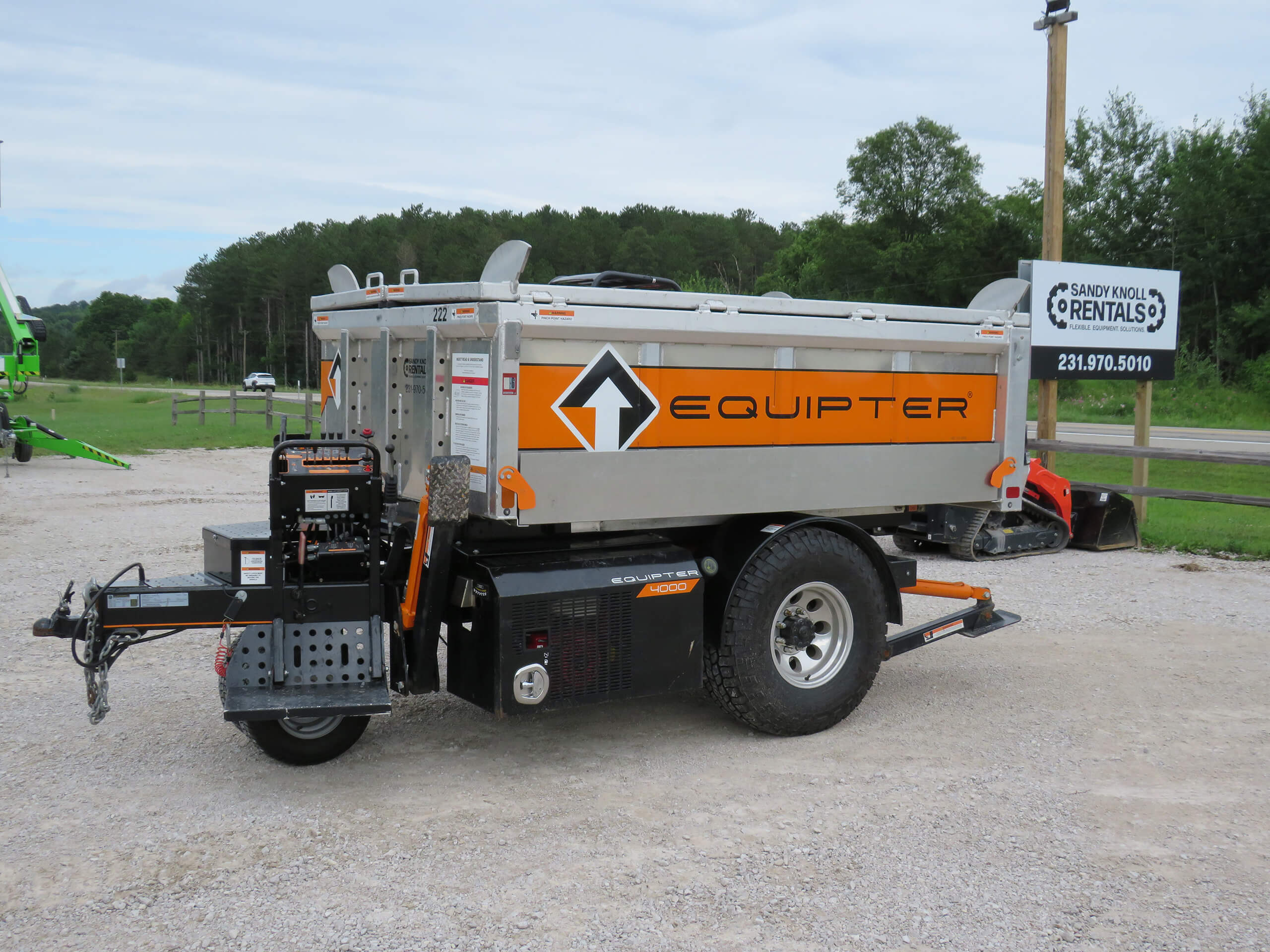 A parked Equipter debris removal trailer with orange branding, positioned on a gravel lot near a Sandy Knoll Rentals signpost and trees in the background.
