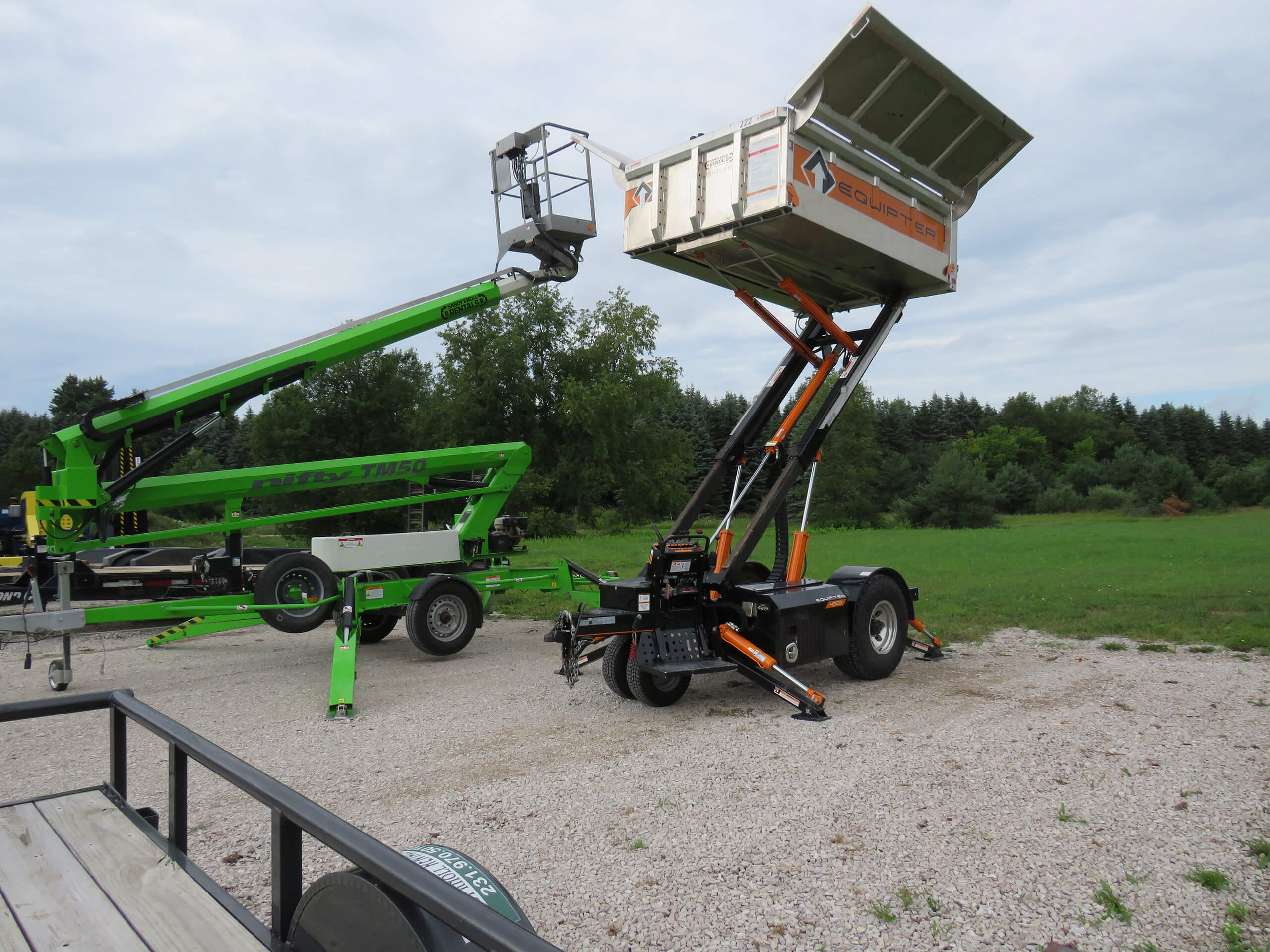 A green boom lift and a trailer with an elevated dump bed are parked on a gravel area near a grassy field with trees in the background.