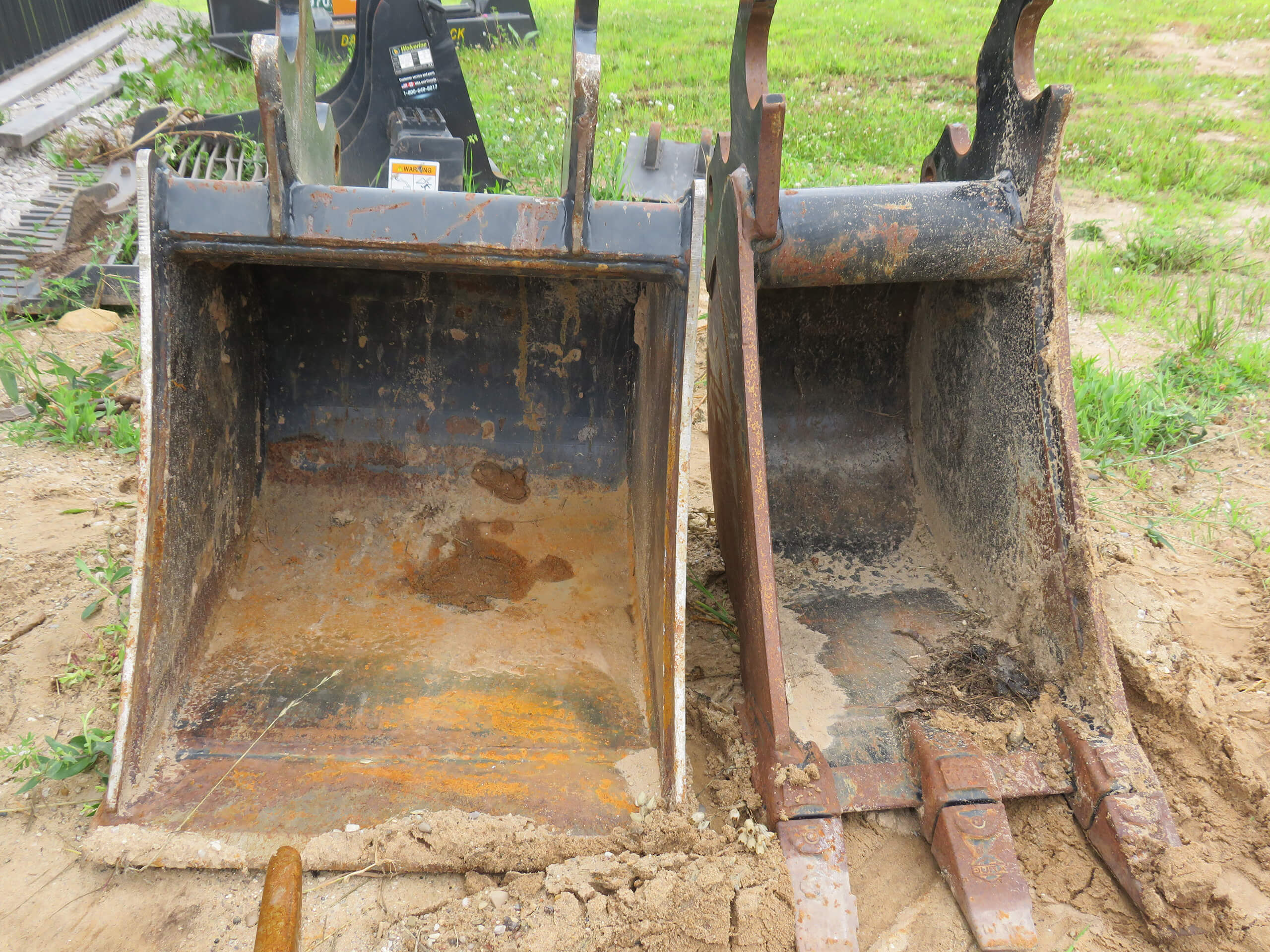 Two rusty, worn excavator buckets are positioned side by side on a dirt surface, with grass and construction equipment visible in the background.