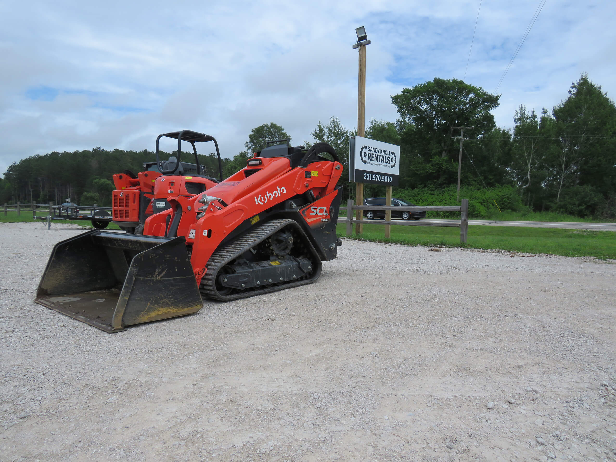 An orange Kubota skid steer loader is parked on a gravel lot near a wooden fence, with a rental sign and green trees in the background.
