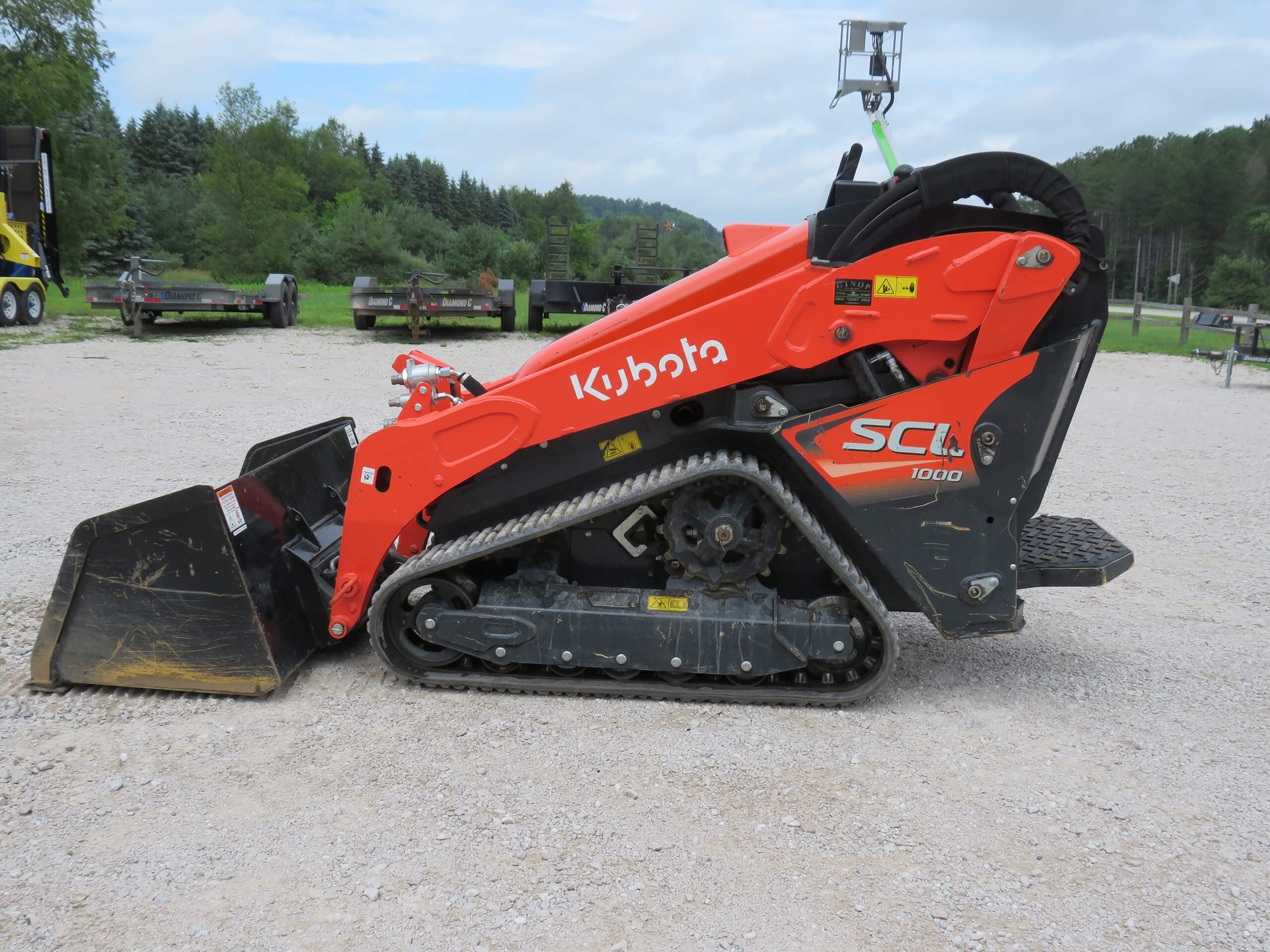 A red Kubota SCL1000 compact track loader with a front bucket attachment parked on gravel, with trees and trailers in the background.
