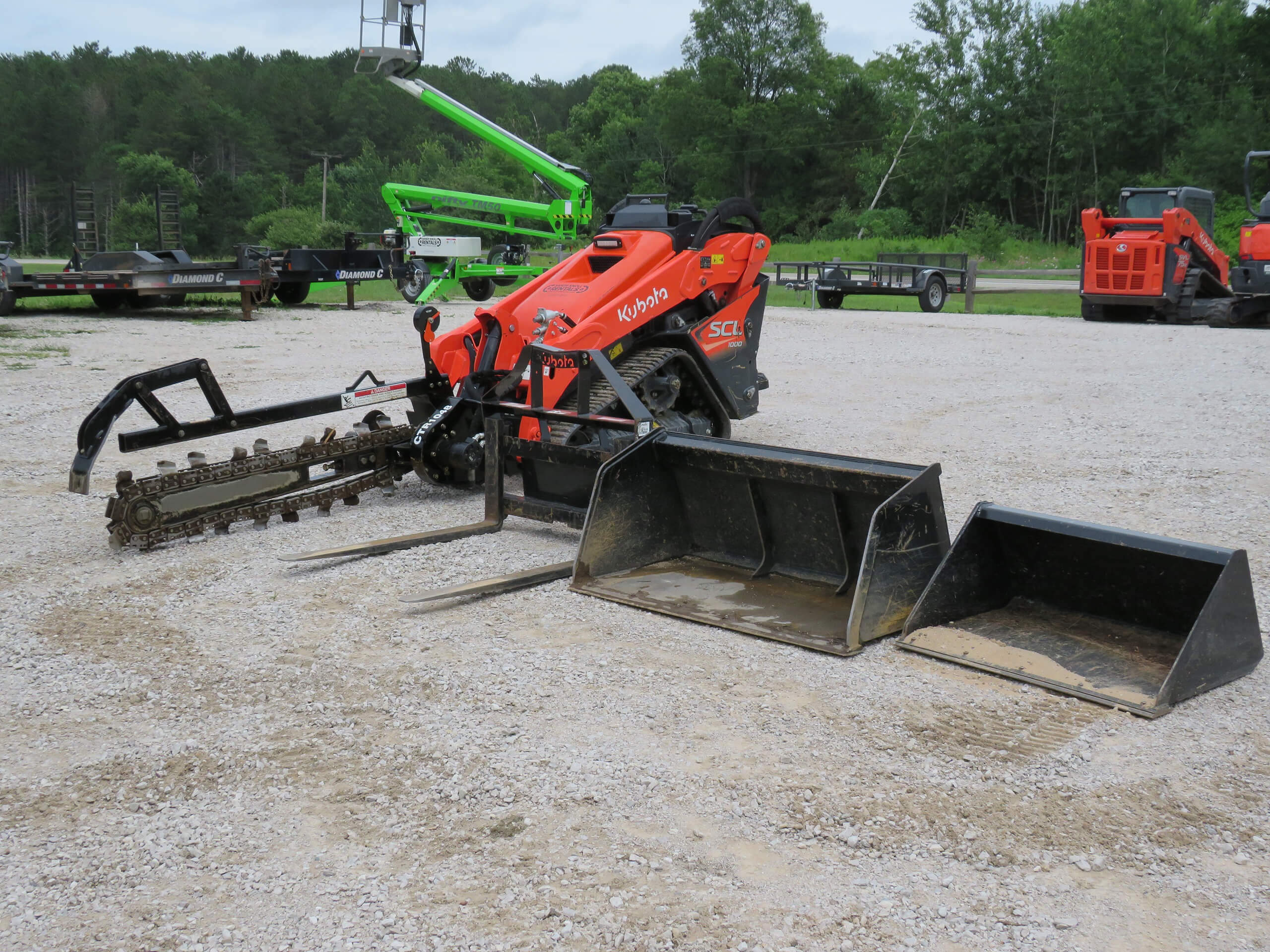An orange Kubota compact utility machine with a trencher attachment and three different buckets on a gravel lot, surrounded by trees and other equipment.