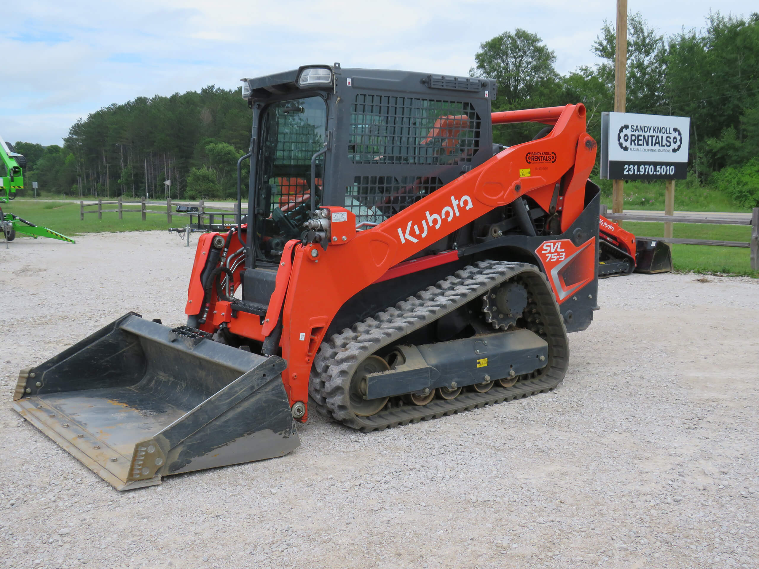A red Kubota SVL75-2 compact track loader is parked on a gravel lot near a fence and a sign for equipment rentals.