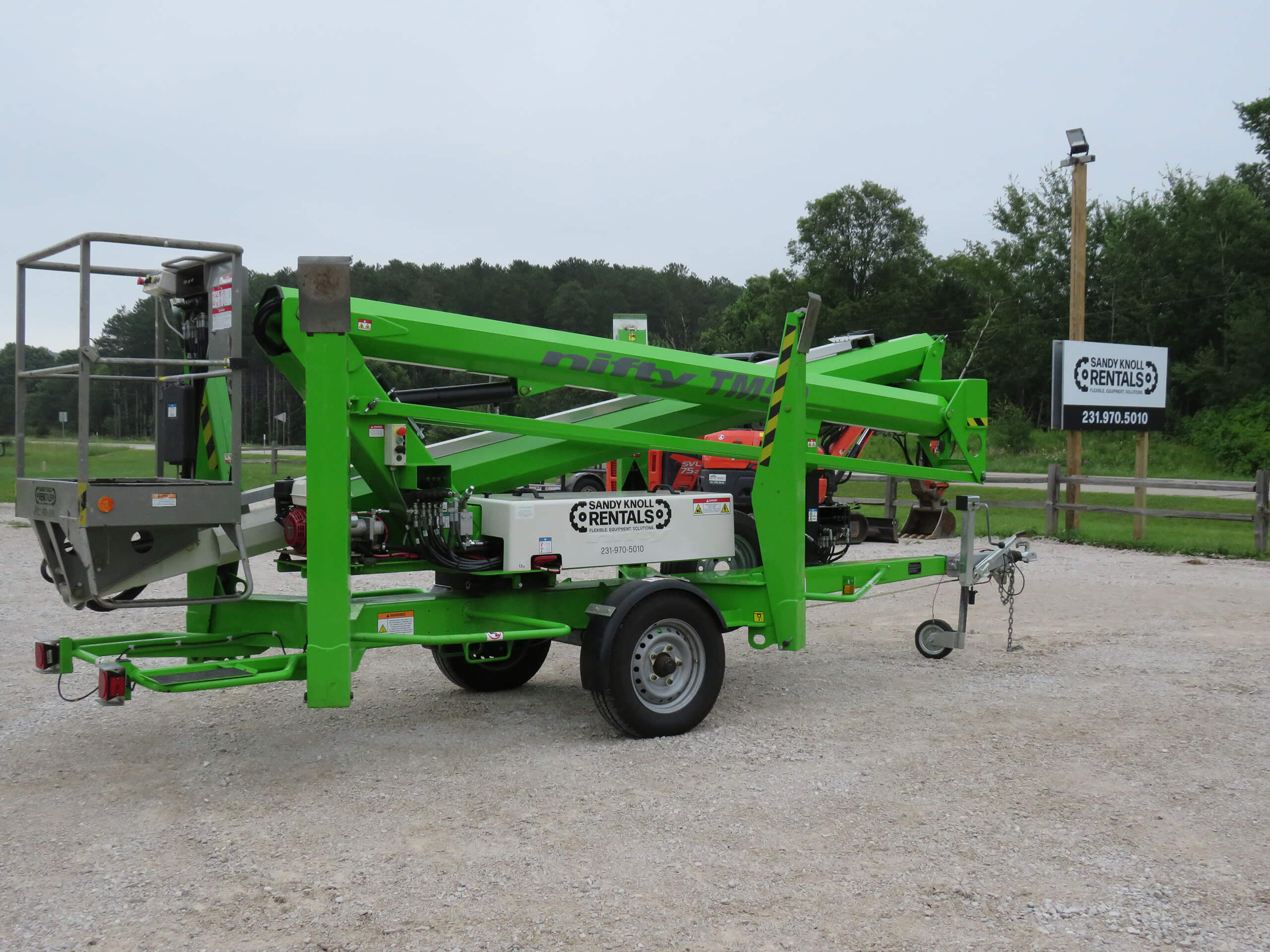 A bright green trailer-mounted boom lift is parked on gravel in front of a wooden fence and trees, with a rental sign visible in the background.