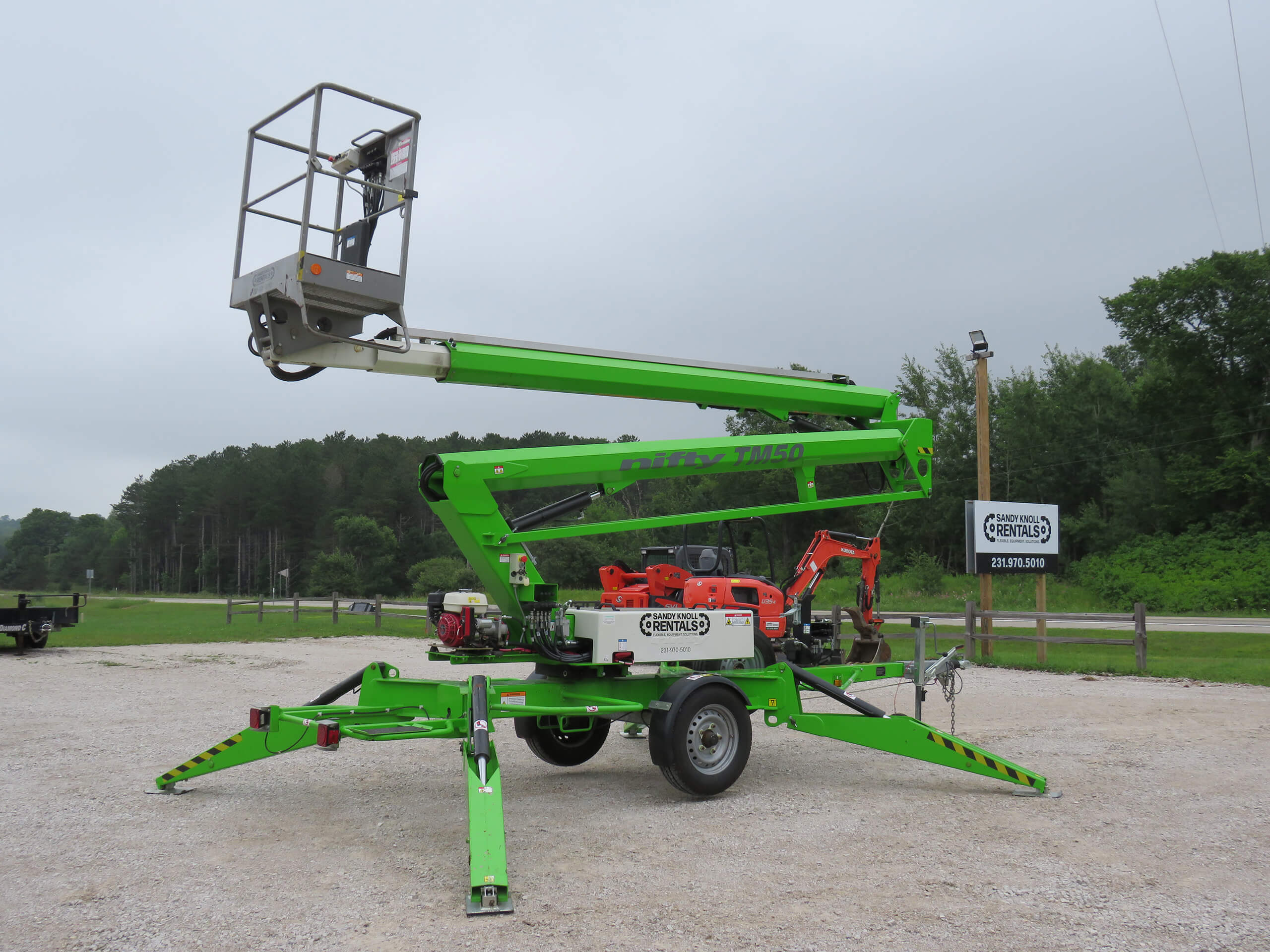 A green towable boom lift with extended arms and stabilizing outriggers is parked on a gravel lot near trees and a wooden fence.