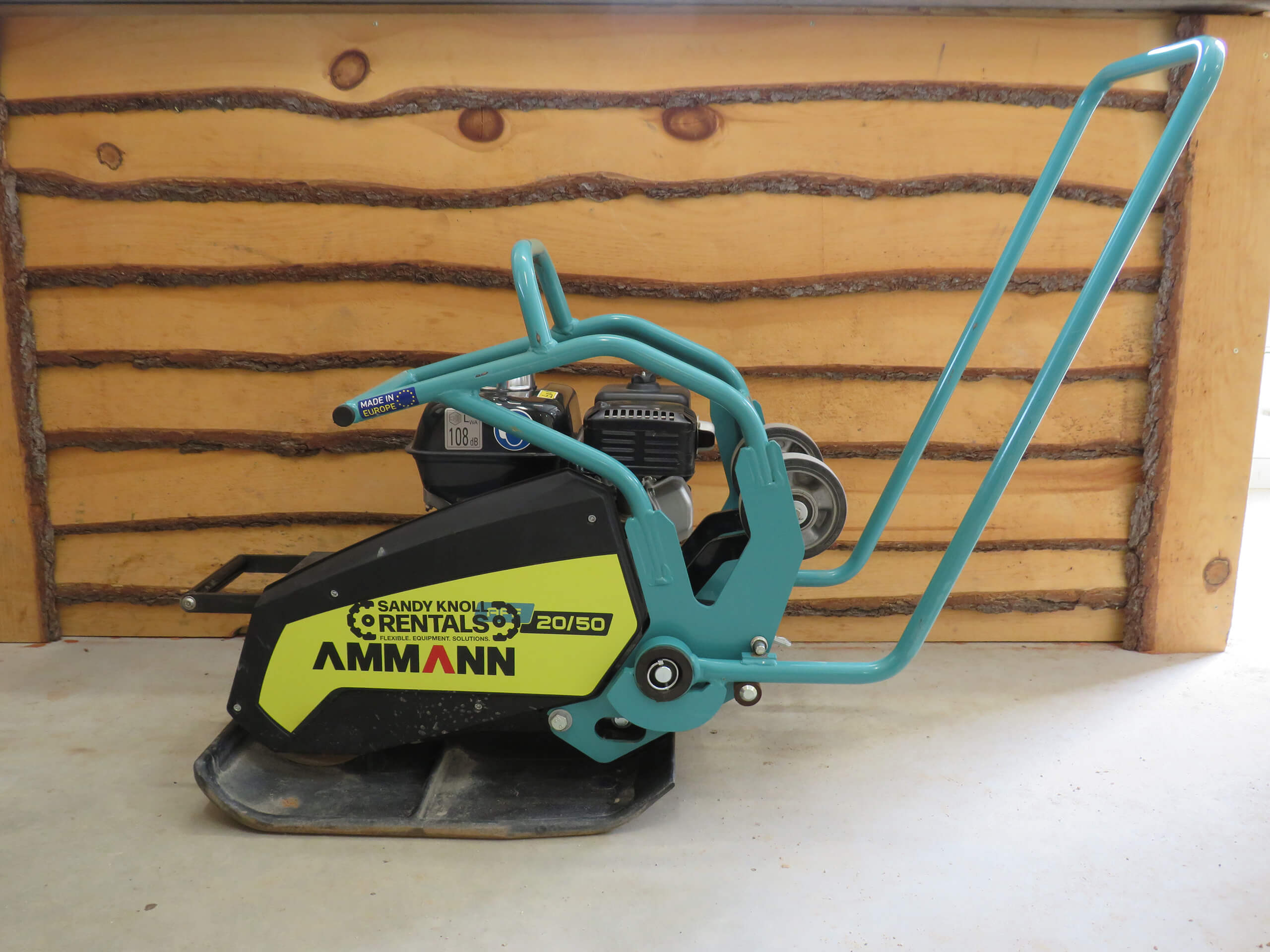 A black and yellow Ammann plate compactor with blue handles, placed indoors in front of a wooden wall.
